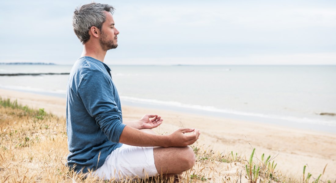 Man meditating on a beach with a calm sea and sky in the background.