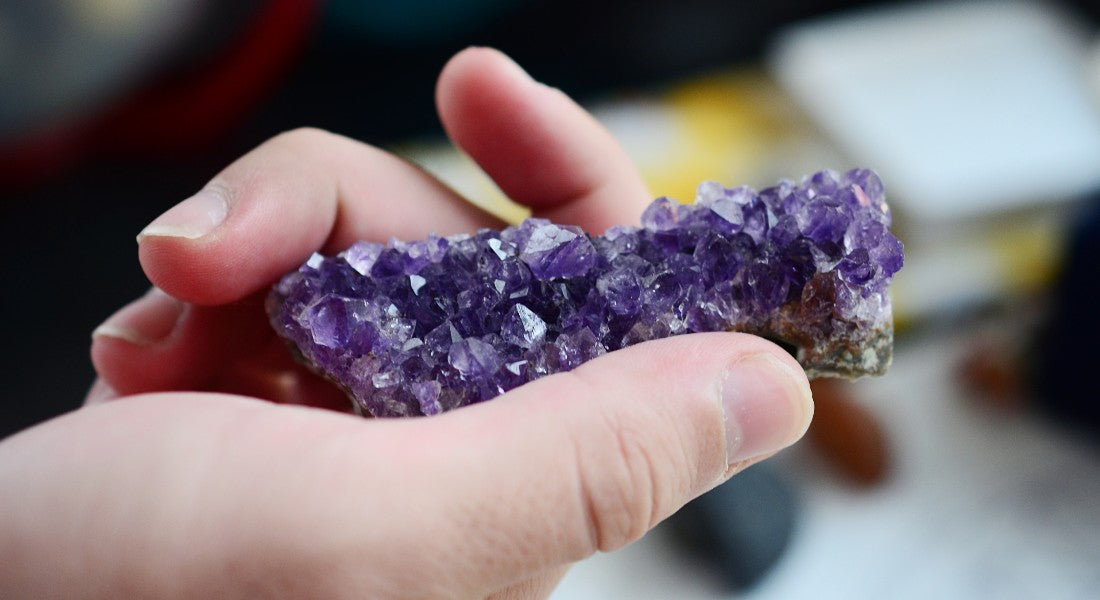 Hand holding a purple amethyst crystal against a blurred background.