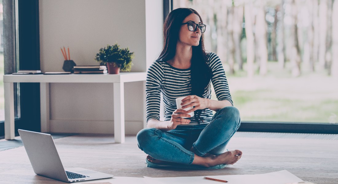Woman sitting cross-legged on the floor with a laptop and notebook, holding a coffee cup.