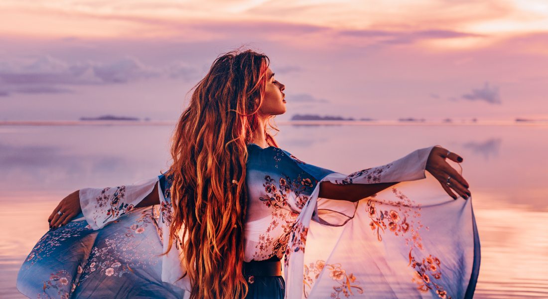 Woman with long hair standing by a body of water at sunset, wearing a floral dress.
