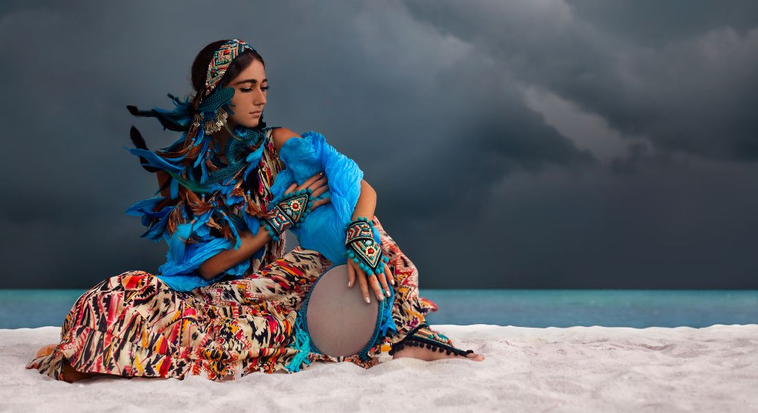 A woman in vibrant shamanic attire sitting on a white sand beach holding a hand drum under a stormy sky.