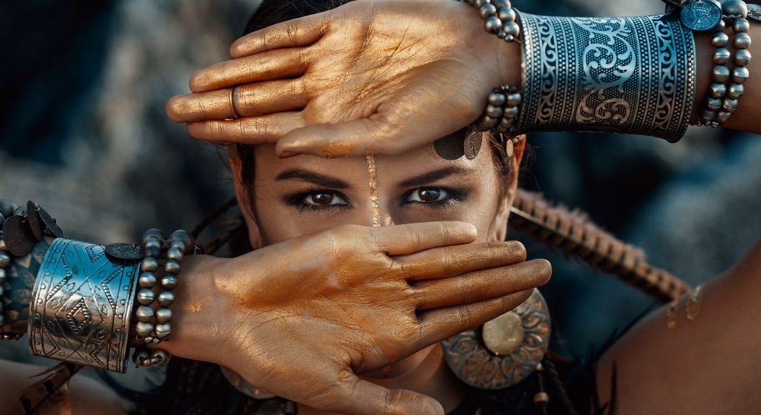Person with bronze hands and face, wearing intricate jewellery against a blurred natural background.