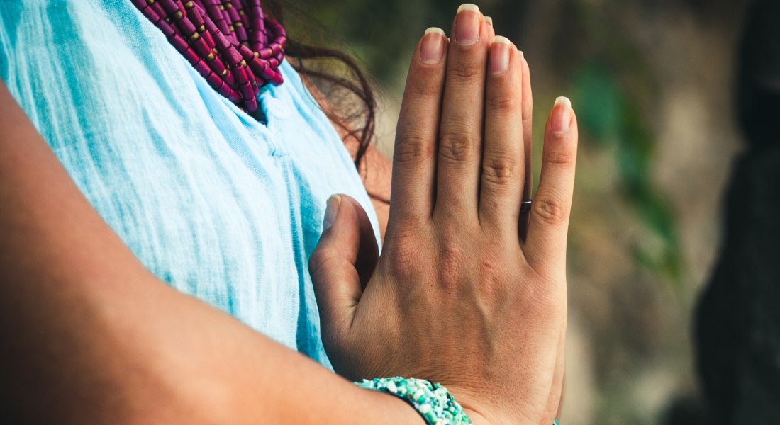 Close-up of a person's hands in a spiritual pose with a blurred natural background.