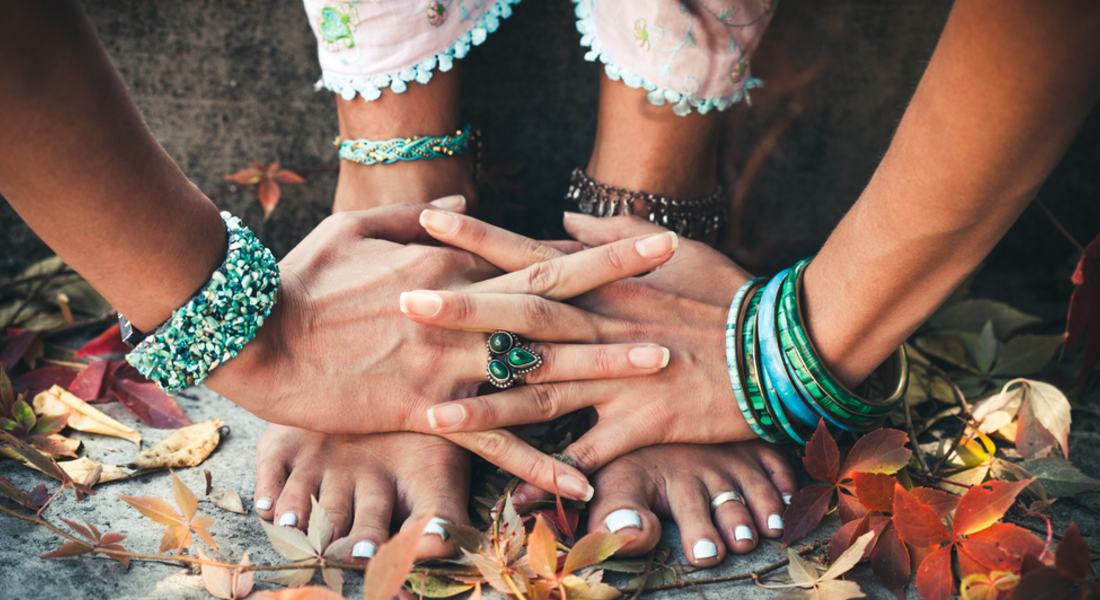 Close-up of hands with green rings and bracelets resting on feet on a natural background.