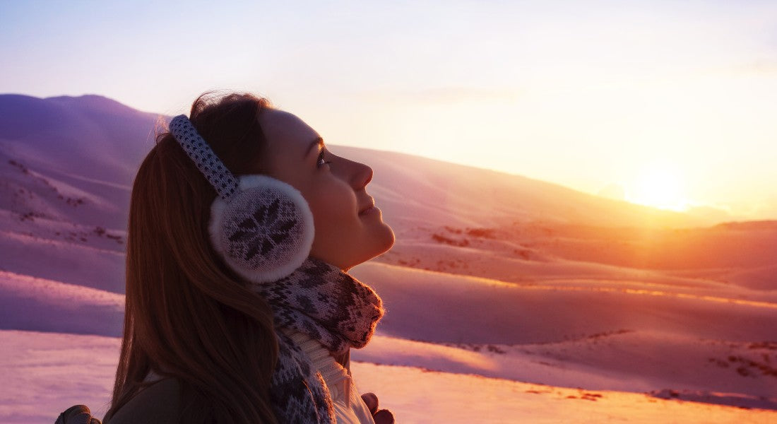 Person wearing winter accessories with a snowy landscape and sunset in the background.