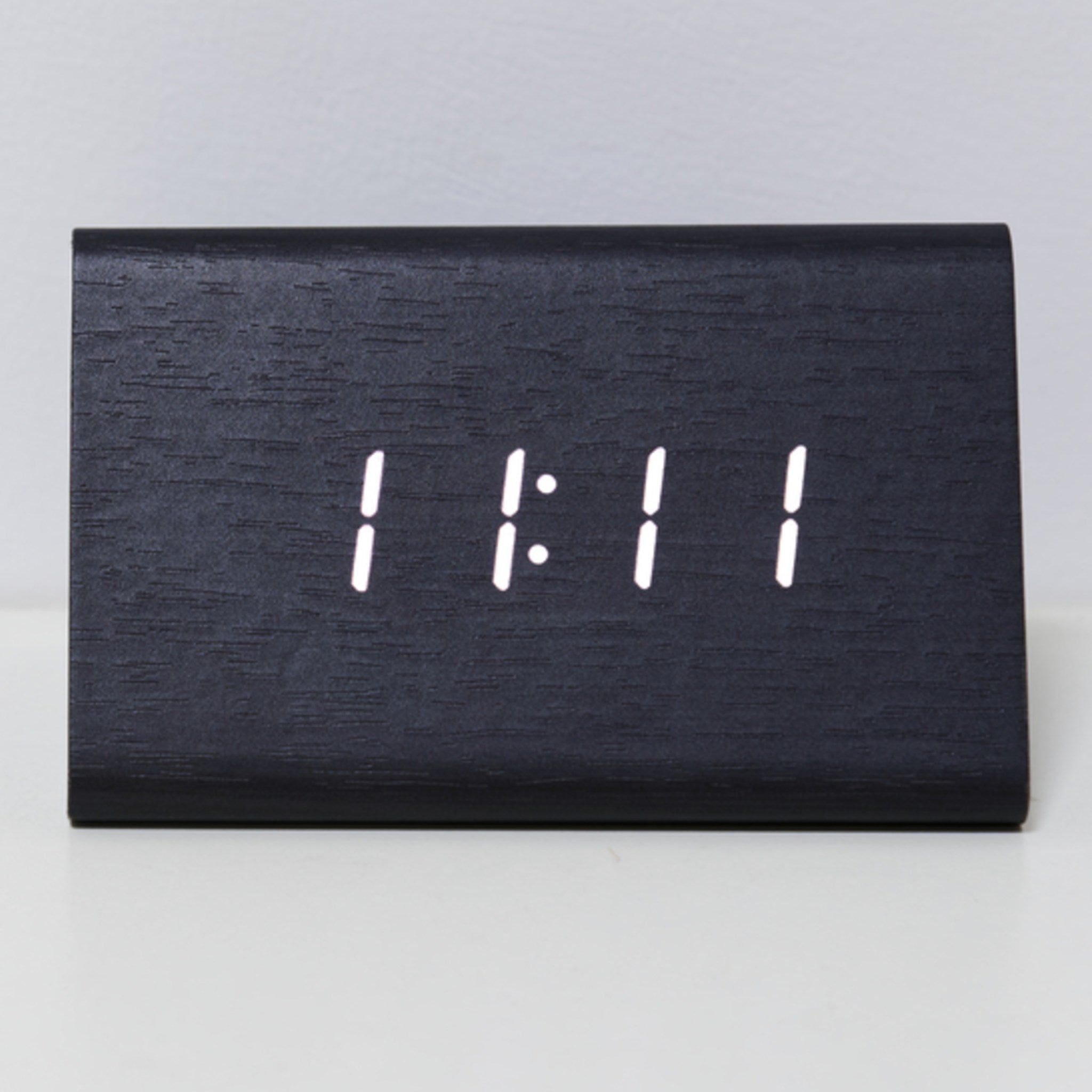 A minimalist black wooden digital clock displaying the number '11:11' in white LED light, resting on a white surface.