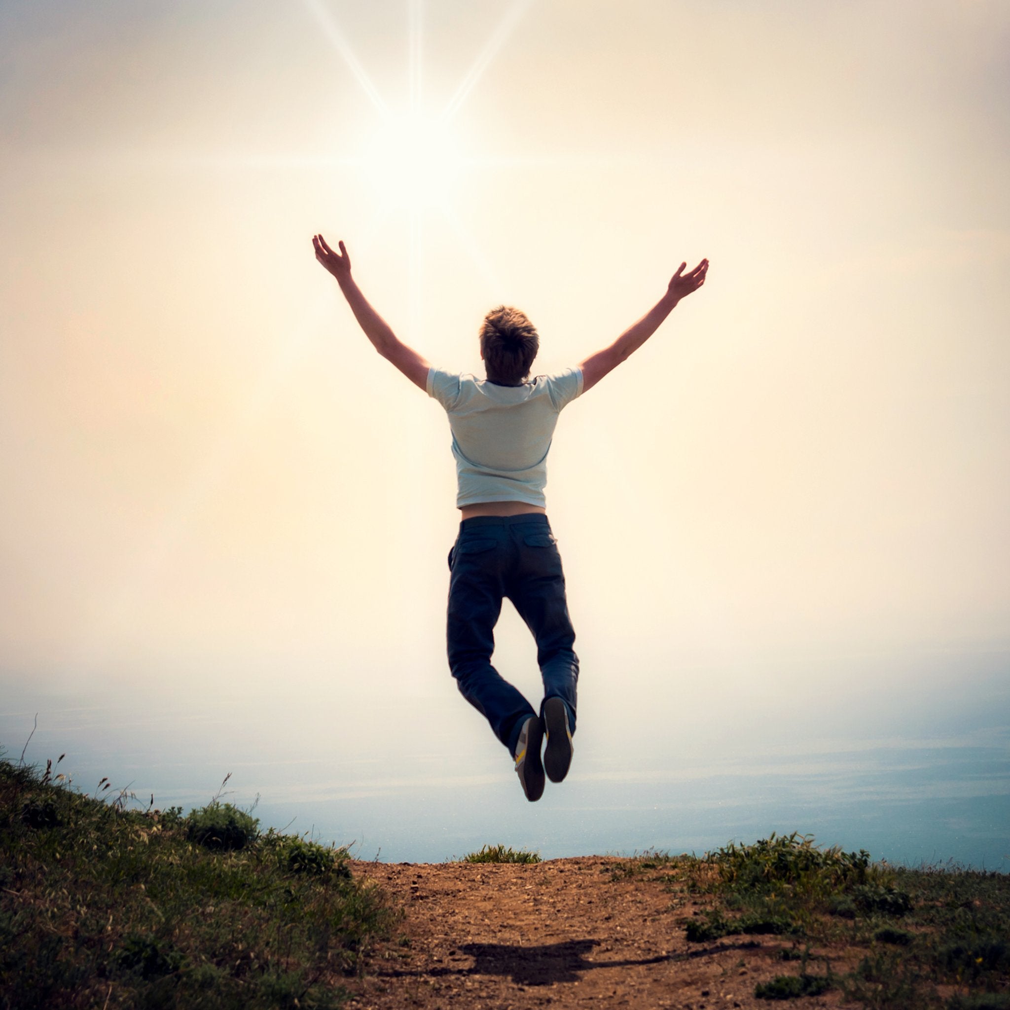 A photograph of a person in a white t-shirt and dark jeans jumping high in the air with their arms outstretched, standing on a dirt path atop a hill. The background is a bright, overexposed sky with a radiating sunburst, symbolising the uplifting energy of the 5555 angel number.