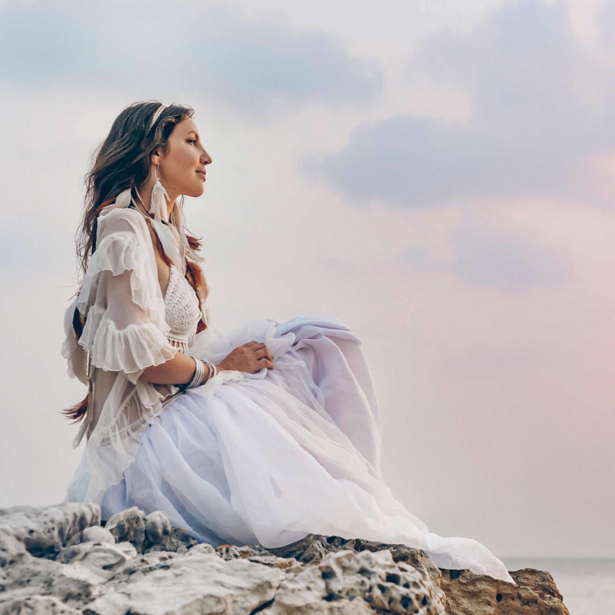 A woman in a flowing white dress and feather accessories sits on a rocky shore, looking towards a pale, sunset-coloured sky, suggesting a spiritual connection to nature and ancestors.