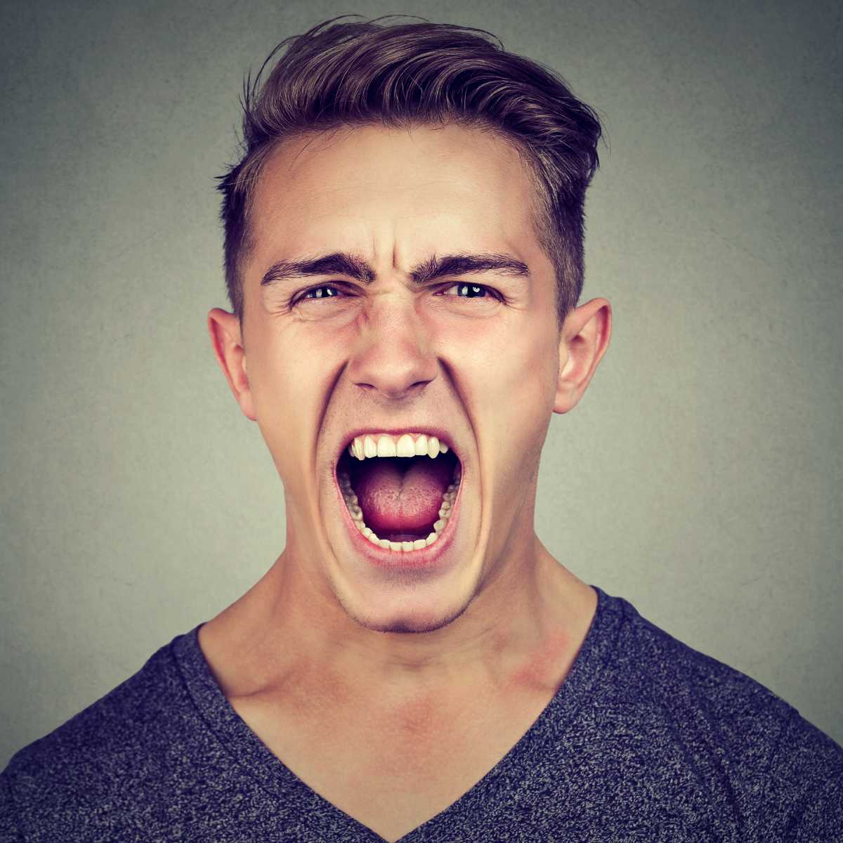 A close-up portrait of a young man shouting with his mouth wide open, wearing a dark heather v-neck t-shirt against a neutral grey background.