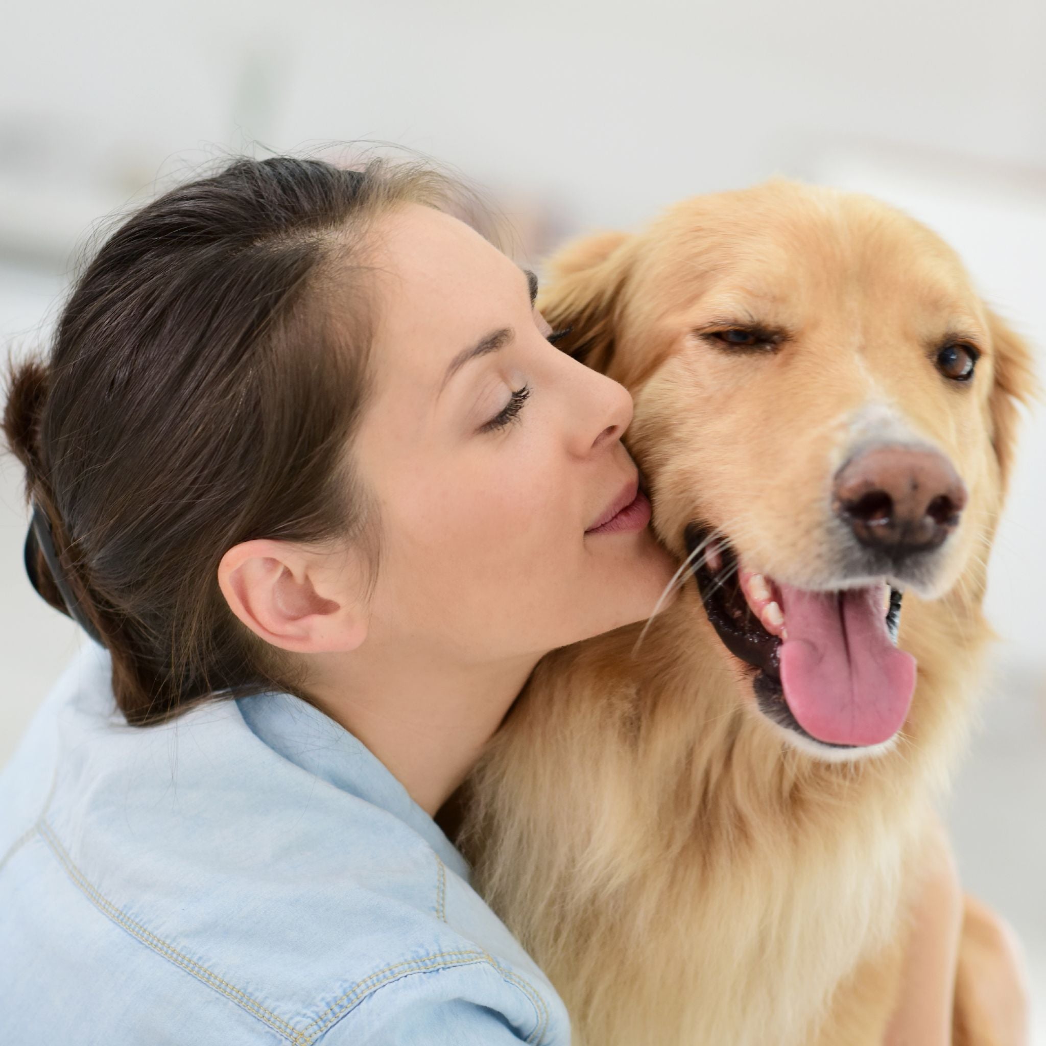 A close-up photograph of a woman hugging a happy golden retriever, gently touching her cheek to the dog's head. The dog has its tongue slightly out and one eye closed in contentment, representing the bond between human and pet and animal healing.