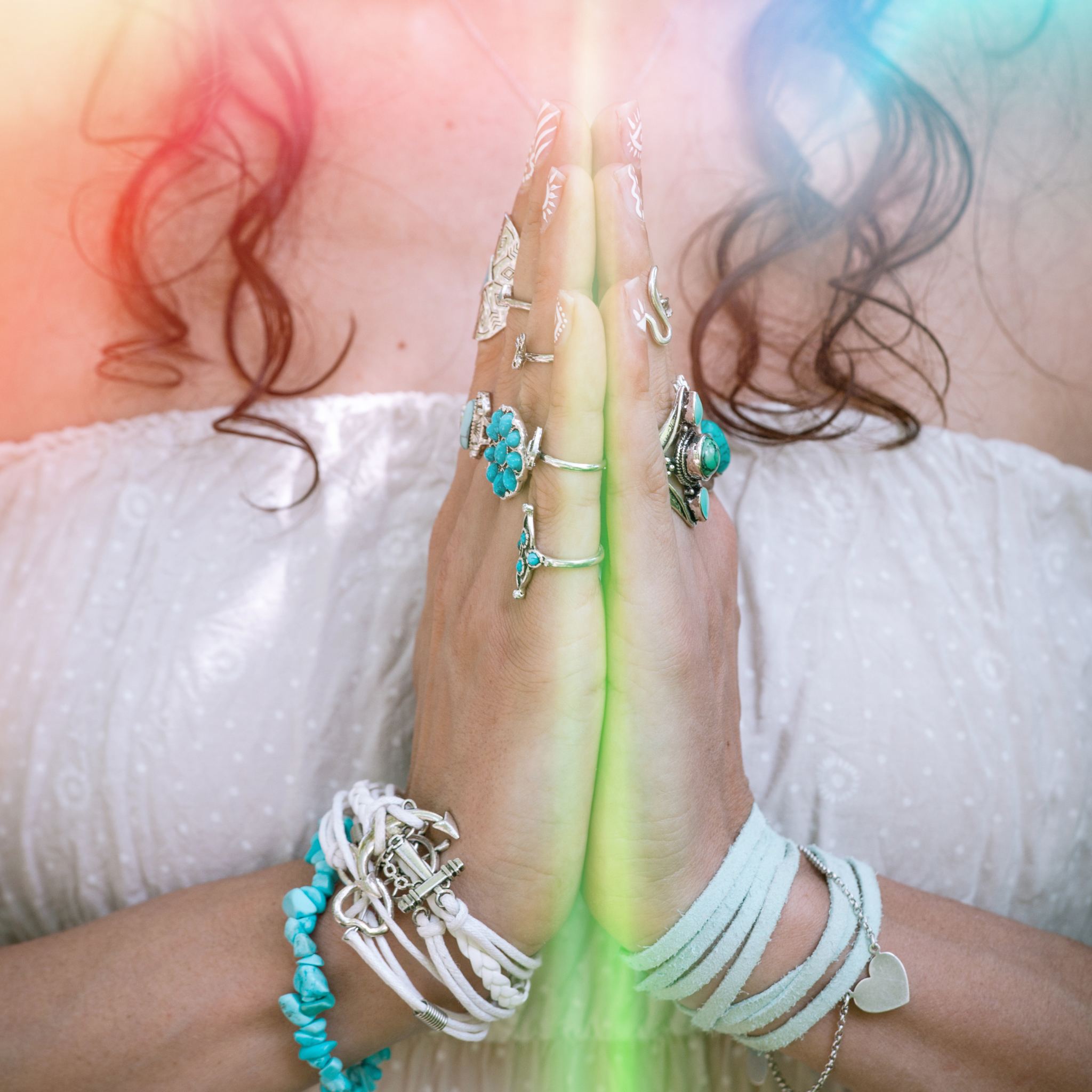 Close-up of a person's hands pressed together in a prayer-like gesture, adorned with rings and bracelets, with soft, colourful light glowing between them. Symbolises Reiki, energy healing, spiritual practice, meditation, and lightwork.