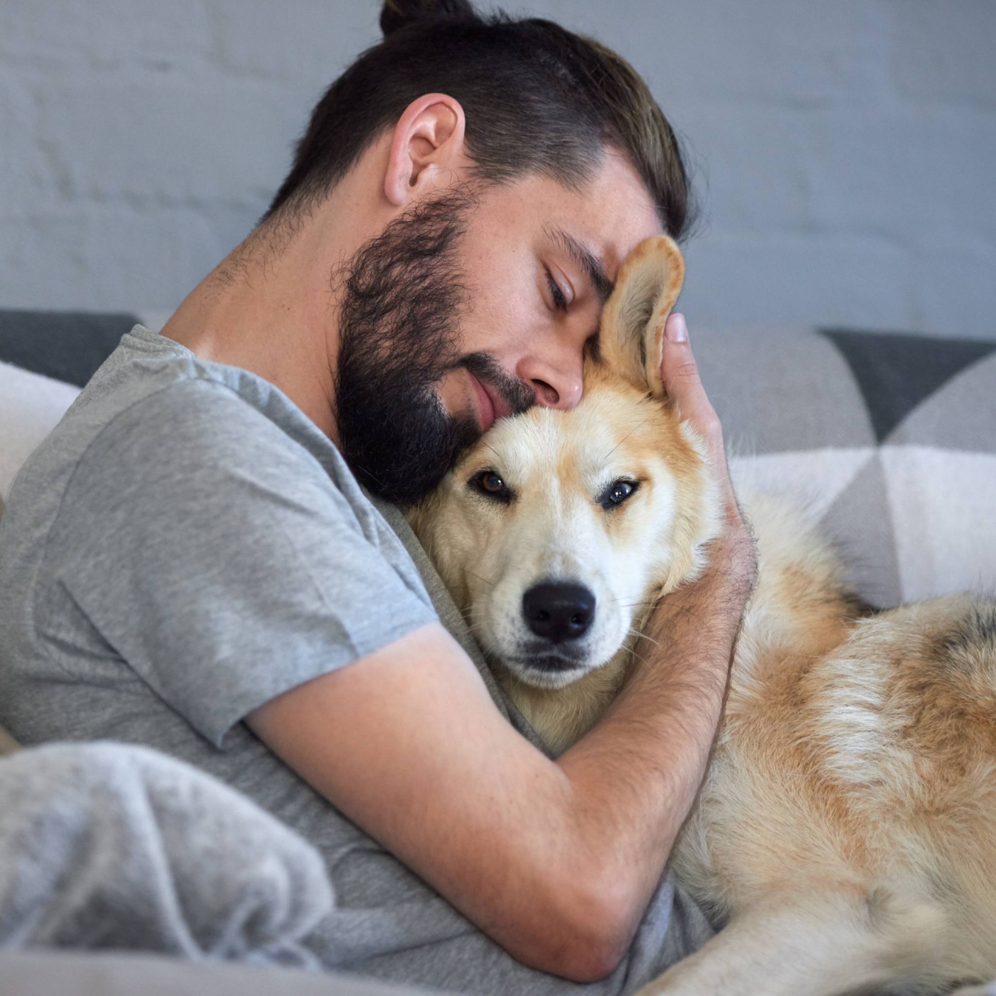 A man with a beard and hair in a bun is seated on a couch, hugging and resting his cheek against a medium-sized, light-coloured dog with one ear slightly folded, while the dog looks directly at the viewer.