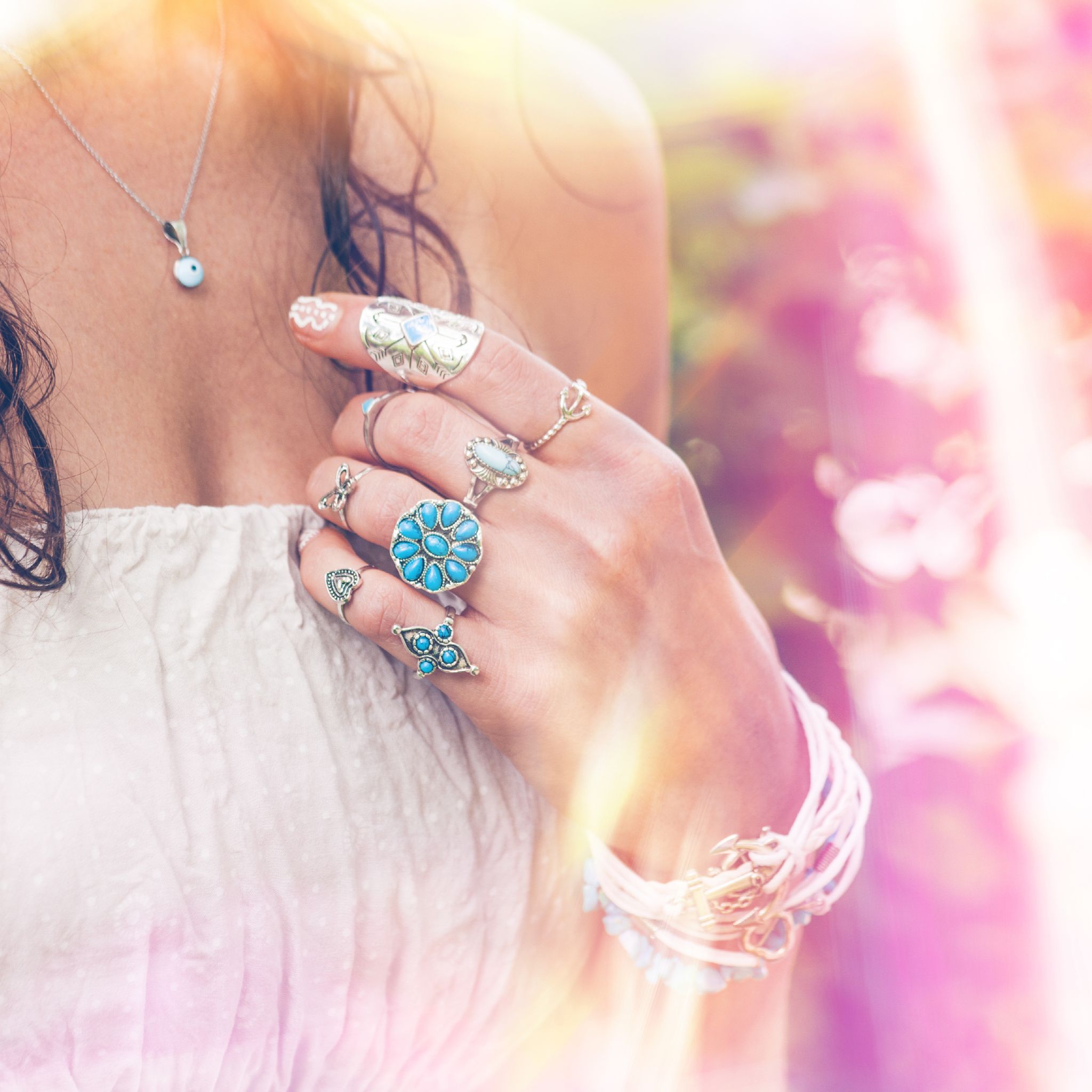 A close-up of a woman's hand adorned with multiple bohemian-style silver and turquoise rings and bracelets, touching her shoulder while standing in soft, bright light that creates pink and golden lens flares.