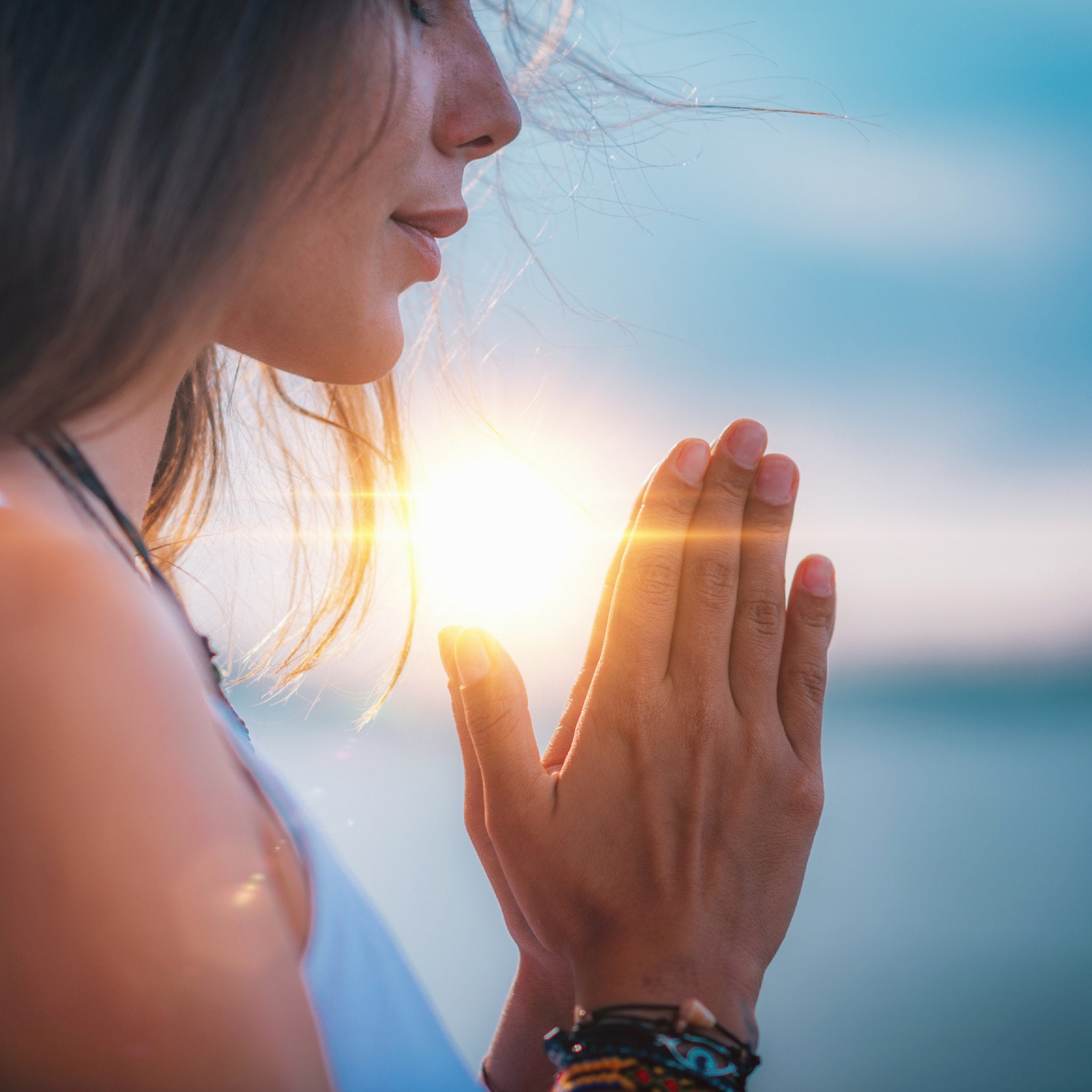 A close-up side profile of a woman meditating outdoors, with her eyes closed and hands held in prayer position (anjali mudra), illuminated by a bright sun flare between her palms against a blurred sky and water background.
