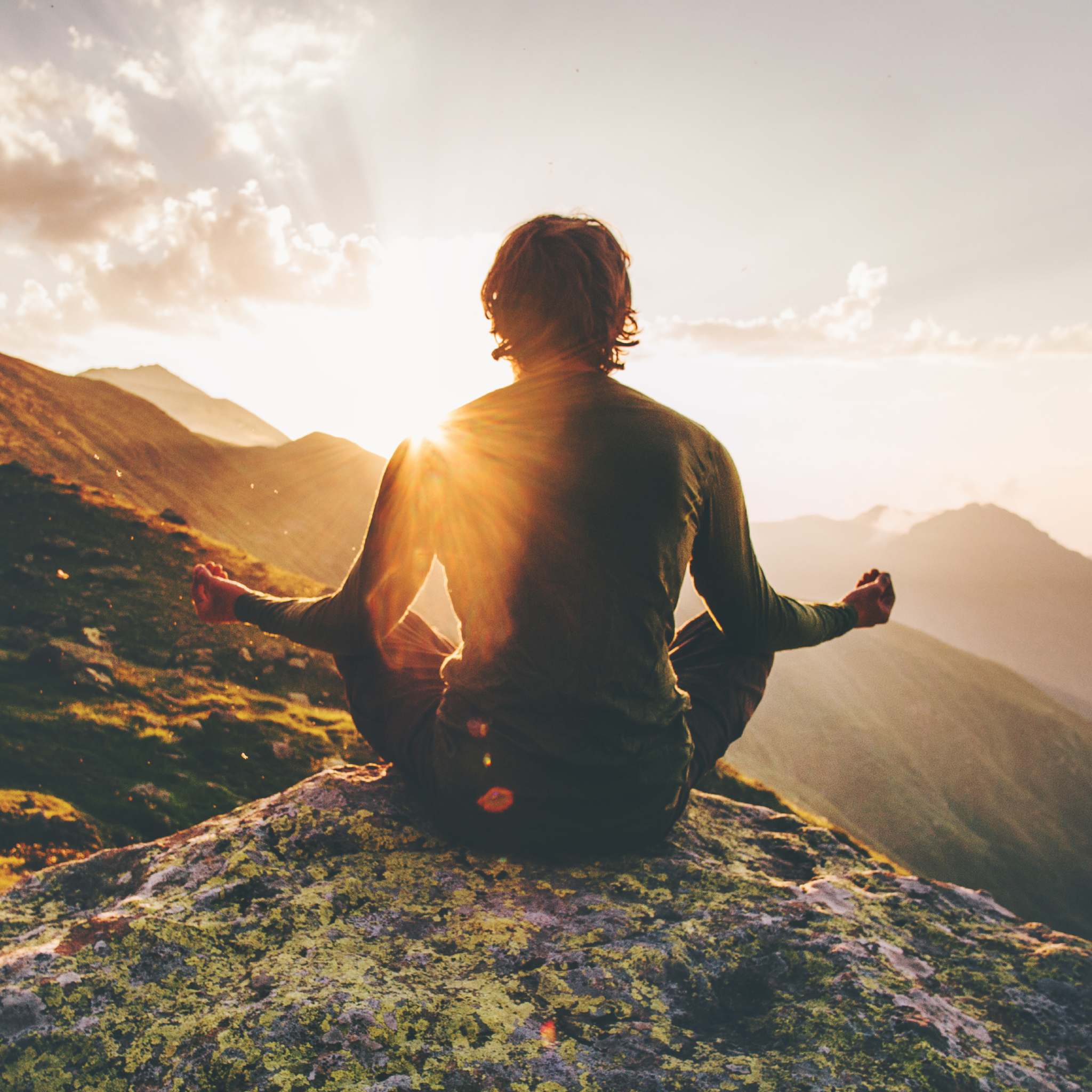 A photograph of a person sitting in the lotus position on a rocky mountaintop, facing away from the camera towards the horizon. The bright, setting sun flares directly behind them, radiating beams of light across the mountains, symbolising ancient wisdom connections.