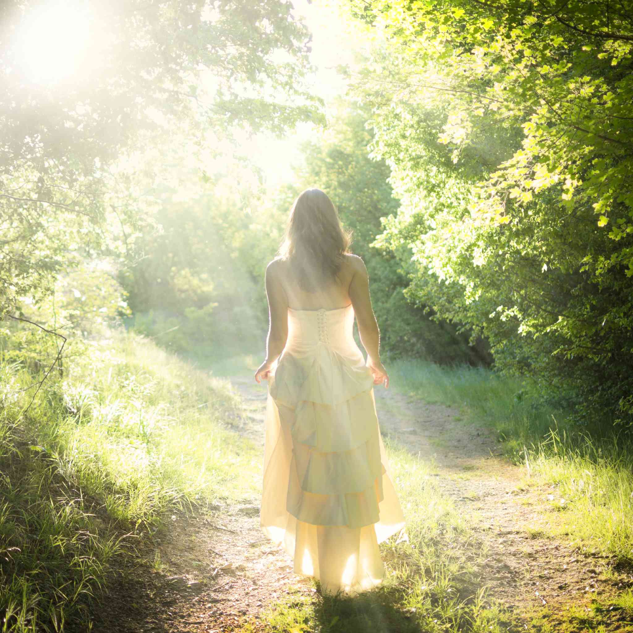 Woman in a long dress walking away on a sunlit forest path representing Aradia.