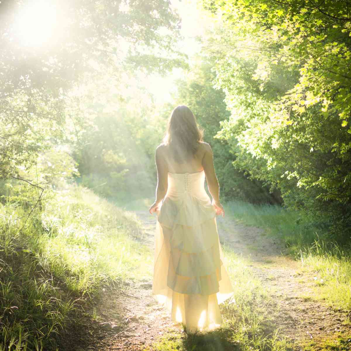 Woman in a long dress walking away on a sunlit forest path representing Aradia.