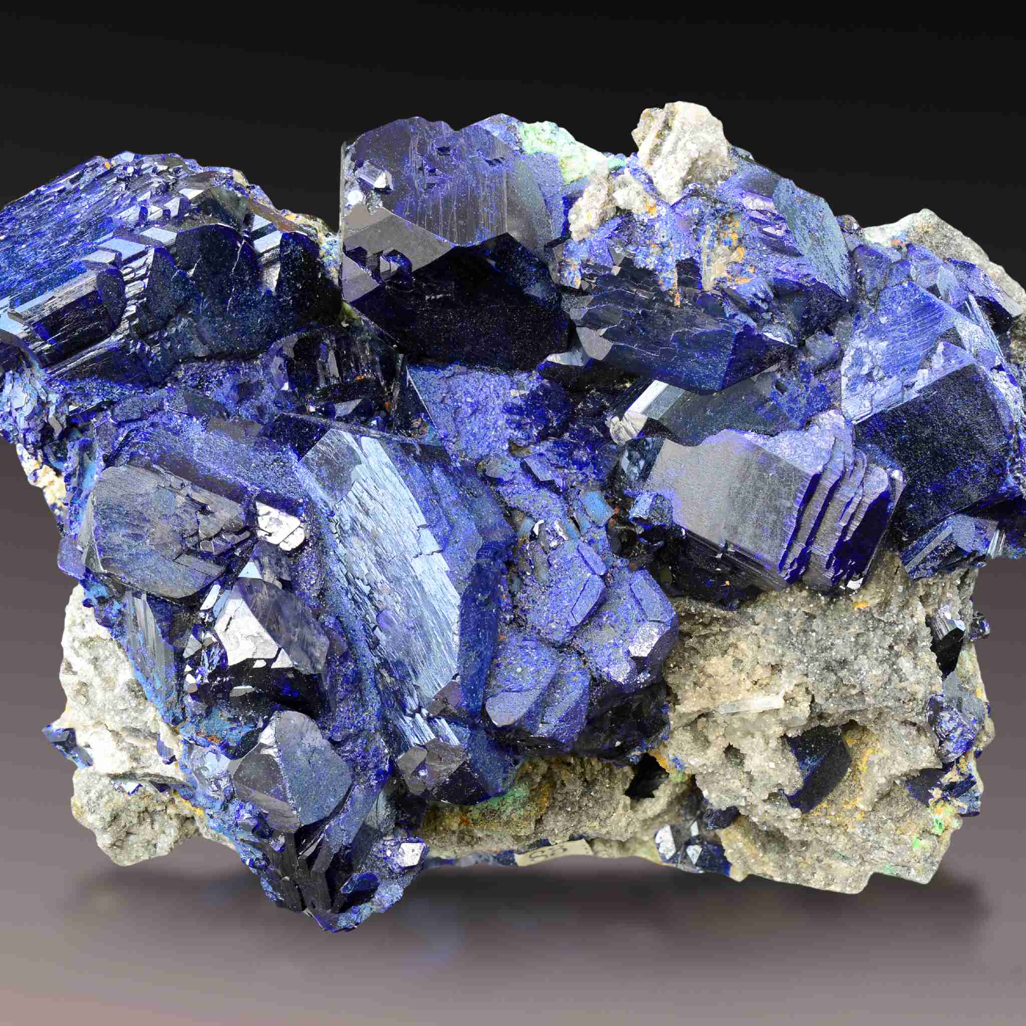 A close-up, high-detail photograph of a dark, crystalline mineral specimen featuring numerous deep blue to indigo facets of azurite crystals, growing on a grey-white rock matrix. The image is set against a dark background, representing the powerful energy of the azurite crystal connections.