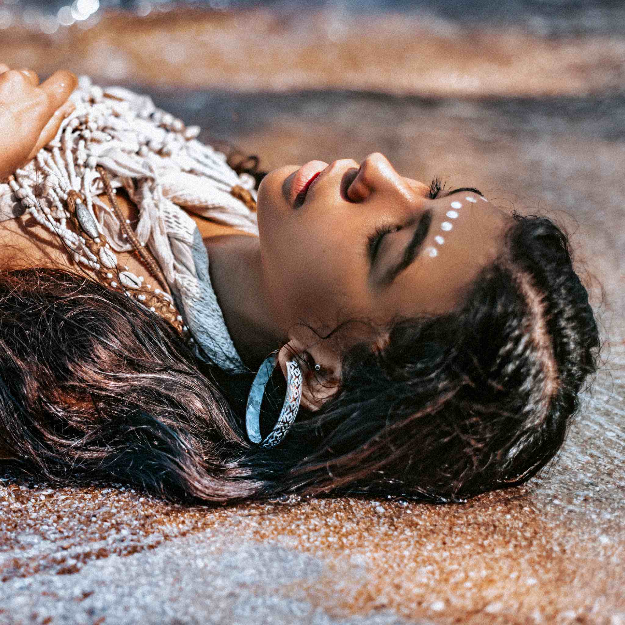 A woman with braids and white dots on her forehead lying on the sand near the water.