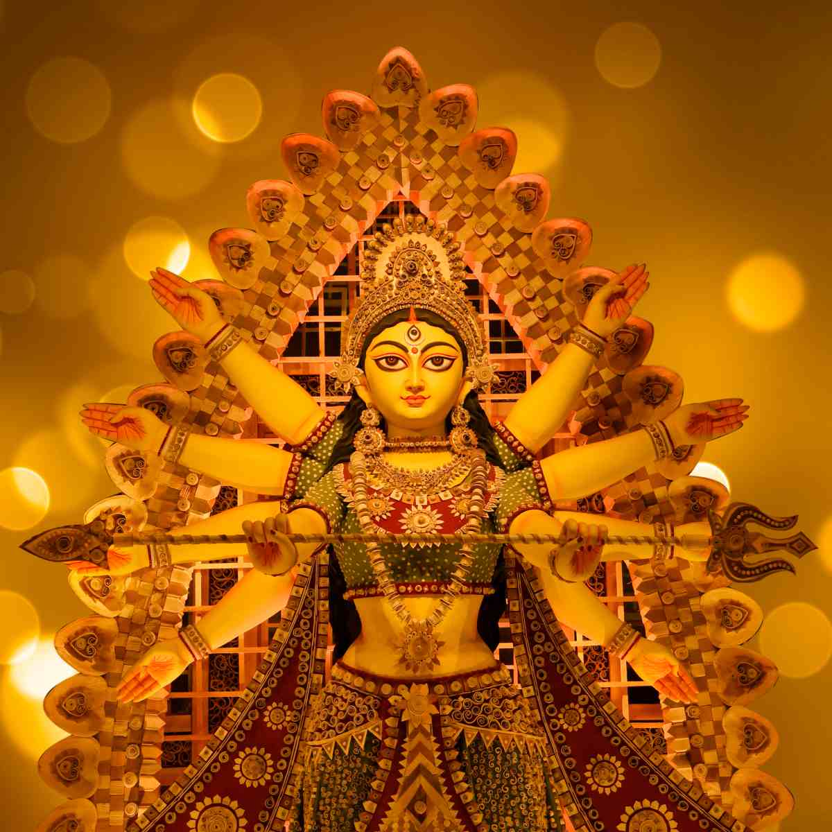 A majestic gold-toned statue of the Hindu Goddess Durga with multiple arms, wearing intricate traditional jewellry and a green attire, set against a warm background of soft, glowing golden bokeh lights.
