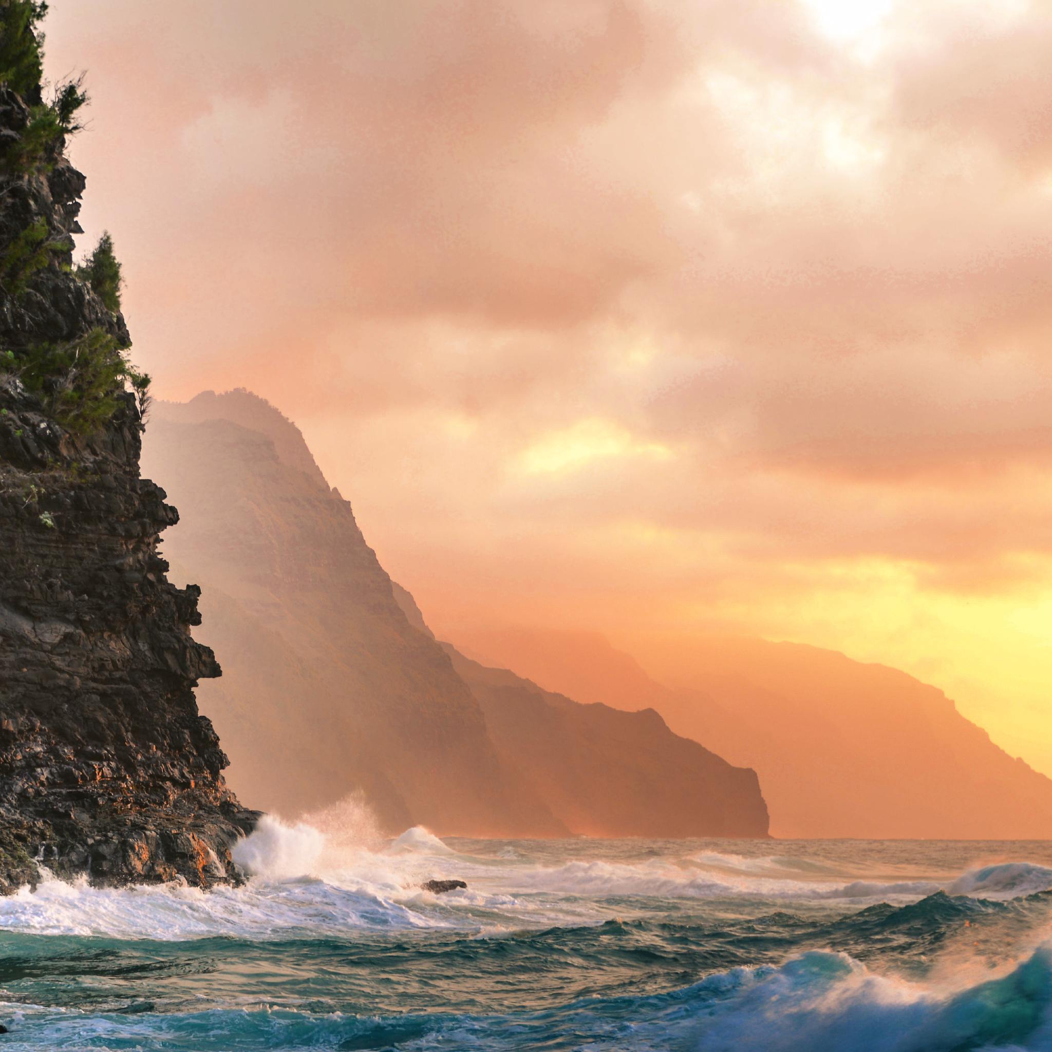 A dramatic coastal landscape at sunset featuring steep, dark rocky cliffs plunging into turquoise ocean waves, set against a glowing sky filled with soft orange and pink clouds.