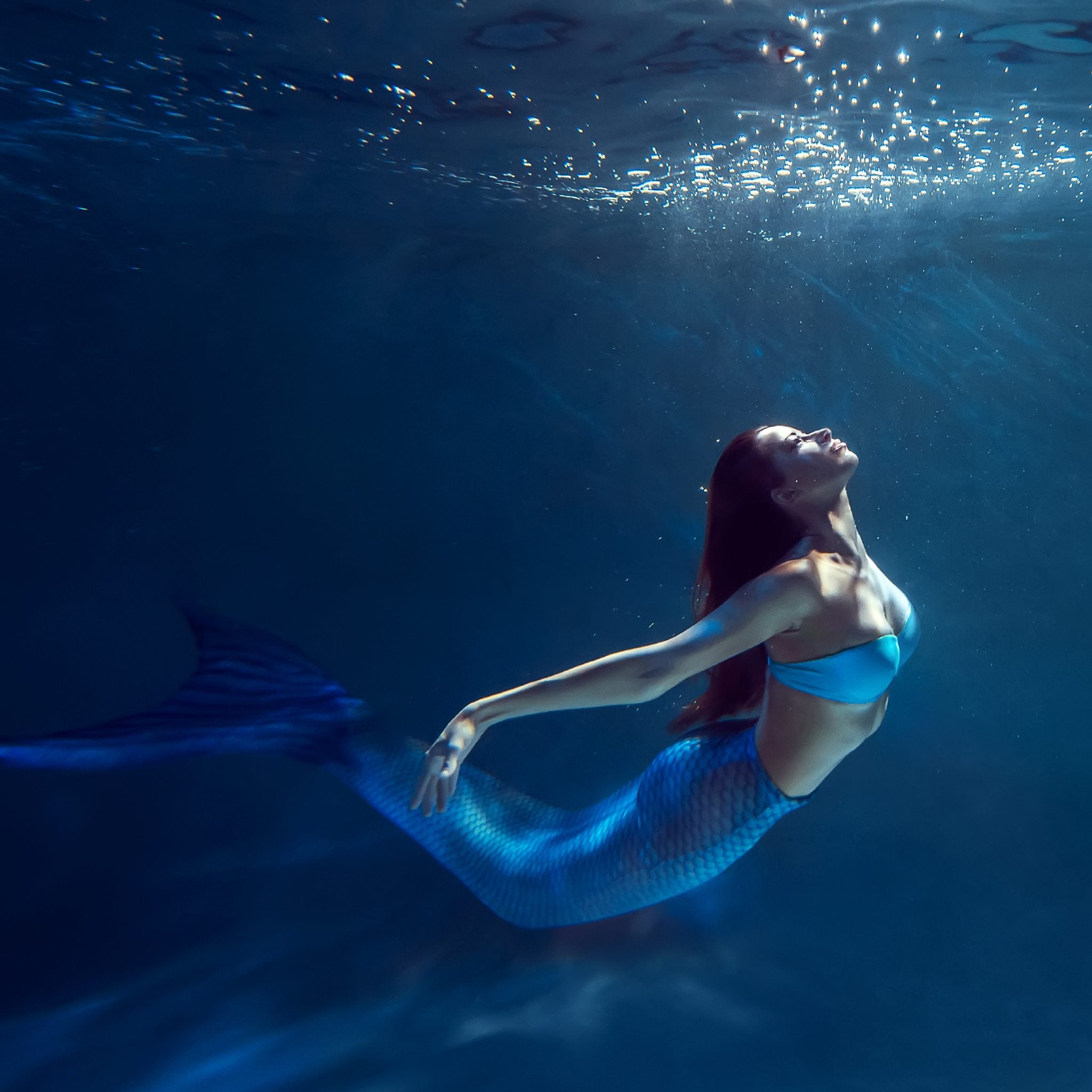 A beautiful woman with long red hair and a shimmery blue mermaid tail swimming gracefully underwater, looking up towards the light, representing mermaid connections.