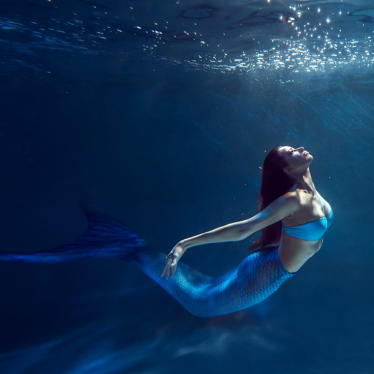 A woman with long red hair and a shimmery blue mermaid tail swimming gracefully underwater.
