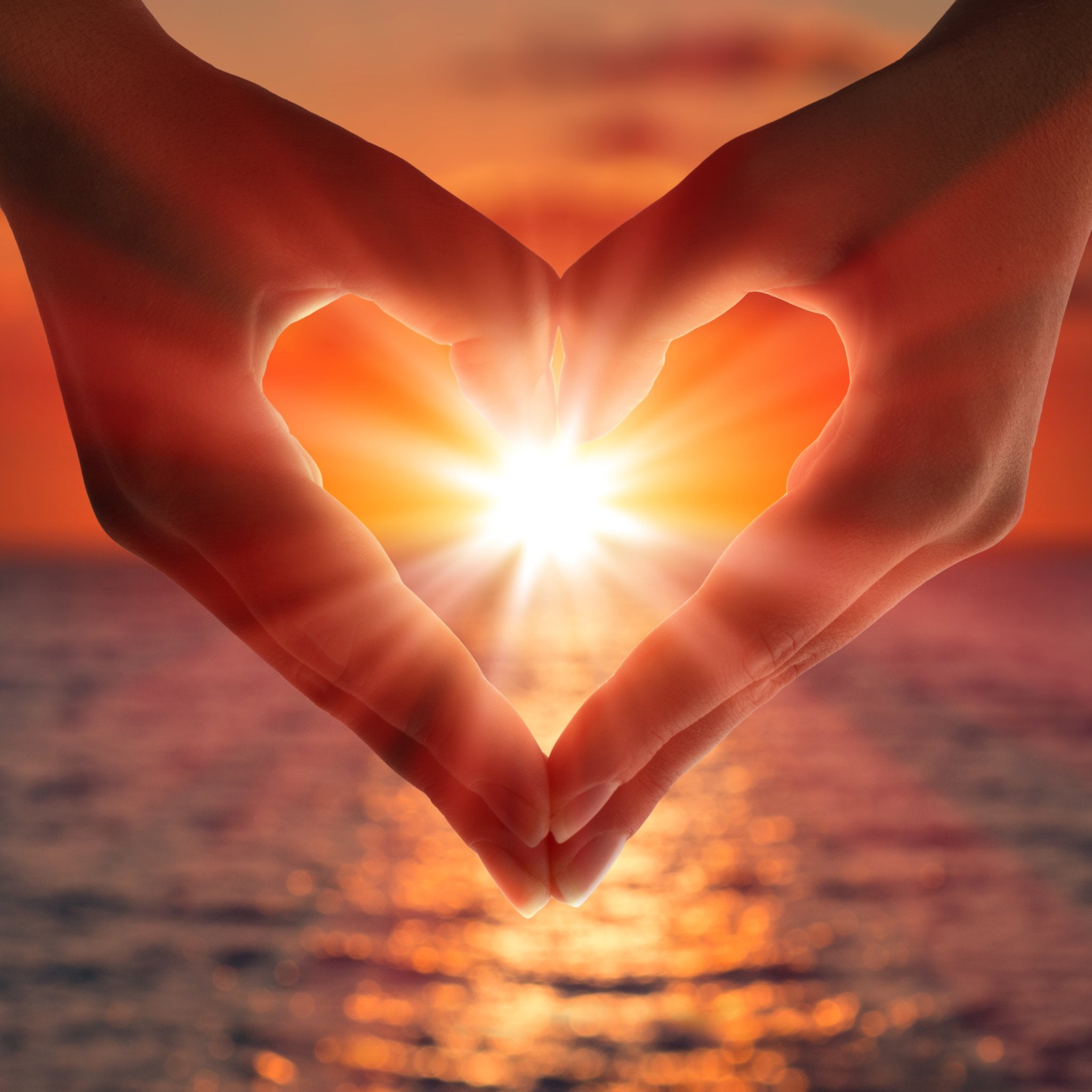 A photograph of a person's hands forming a heart shape with their fingers, framing the bright, setting sun over a shimmering ocean. The warm, fiery orange and red tones convey the energy of dynamic Shakti connections.