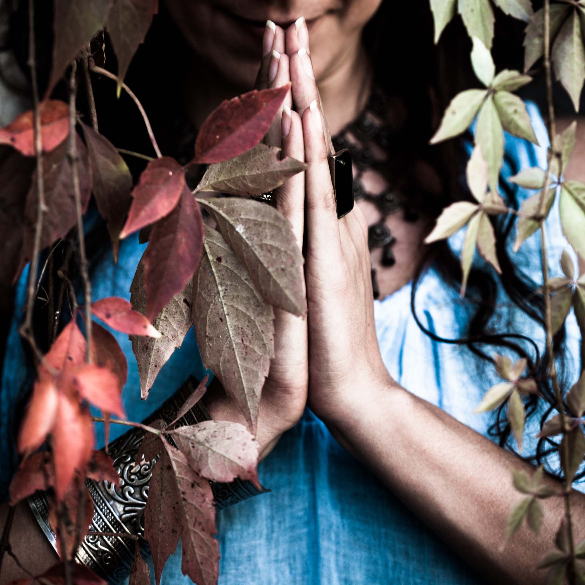 A close-up of a person's hands clasped in a prayer or reverence pose against their chest. They are wearing a flowing blue garment and silver bracelets, framed by red and green autumn leaves. The image conveys a connection with shamanic elders.