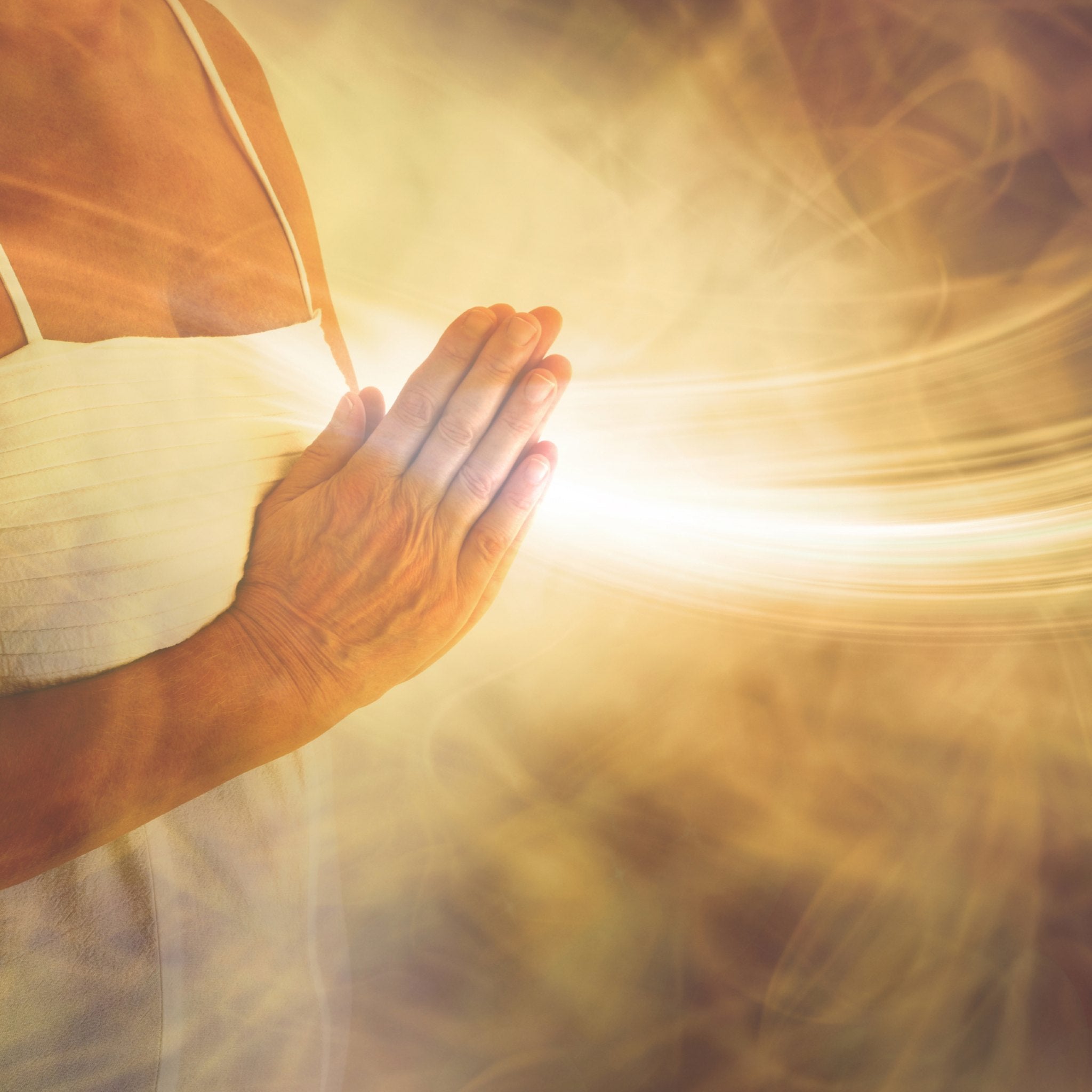 A close-up of a person's torso and hands held in prayer position, with a brilliant, focused stream of golden-yellow light flowing outward from the hands and wrist area against a blurred, swirling, warm-toned background.