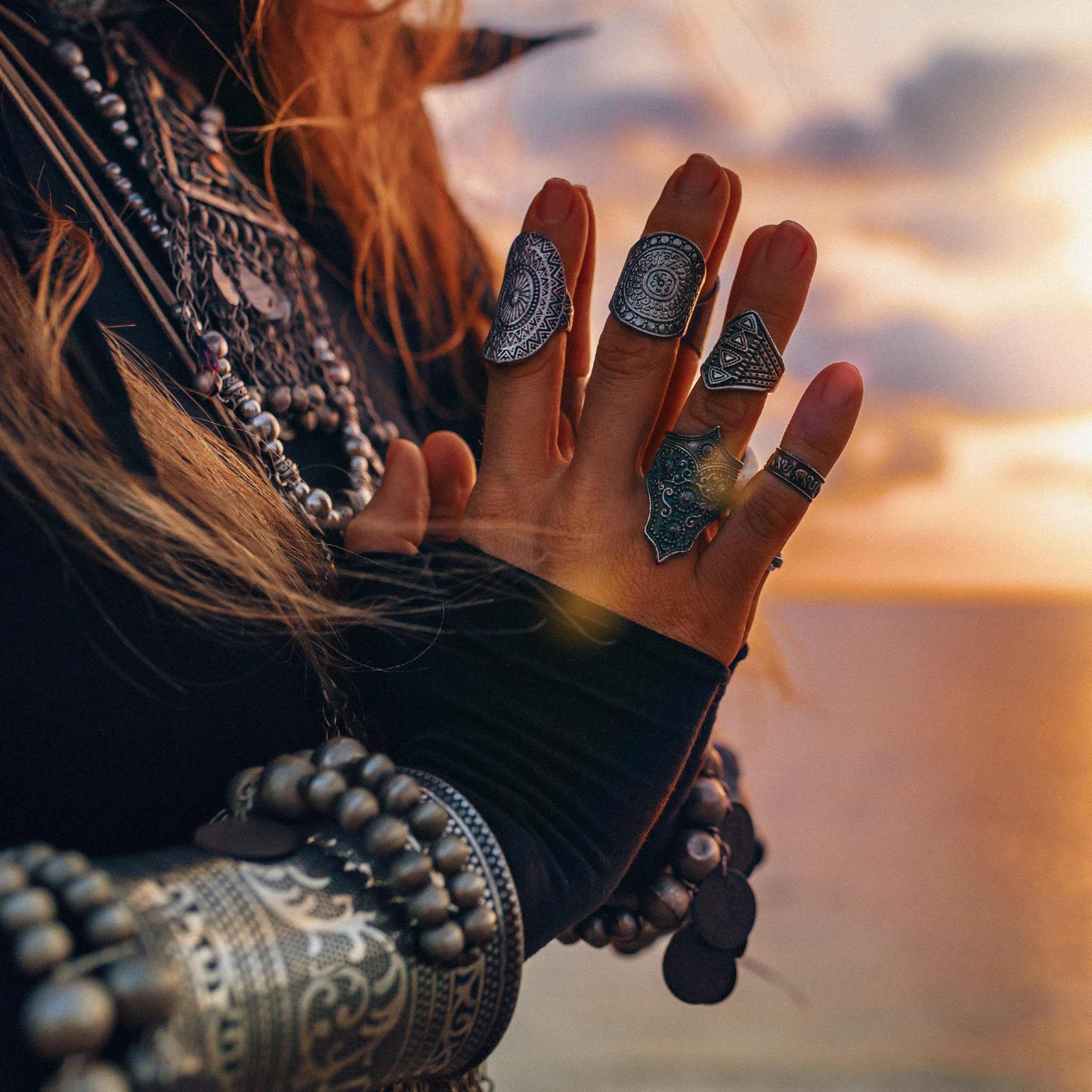 A close-up of a person's hands and arms, heavily adorned with layered silver rings, bracelets, and cuffs, holding their hands in a ritual or contemplative gesture against a warm, sunset-lit sky.