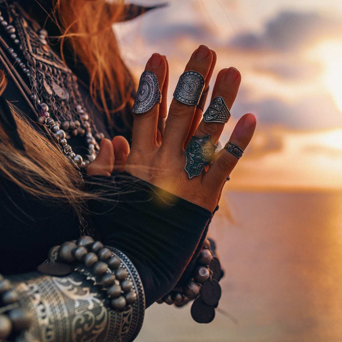 A close-up of a person's hands, heavily adorned with silver rings, bracelets, and cuffs against a warm, sunset-lit sky.