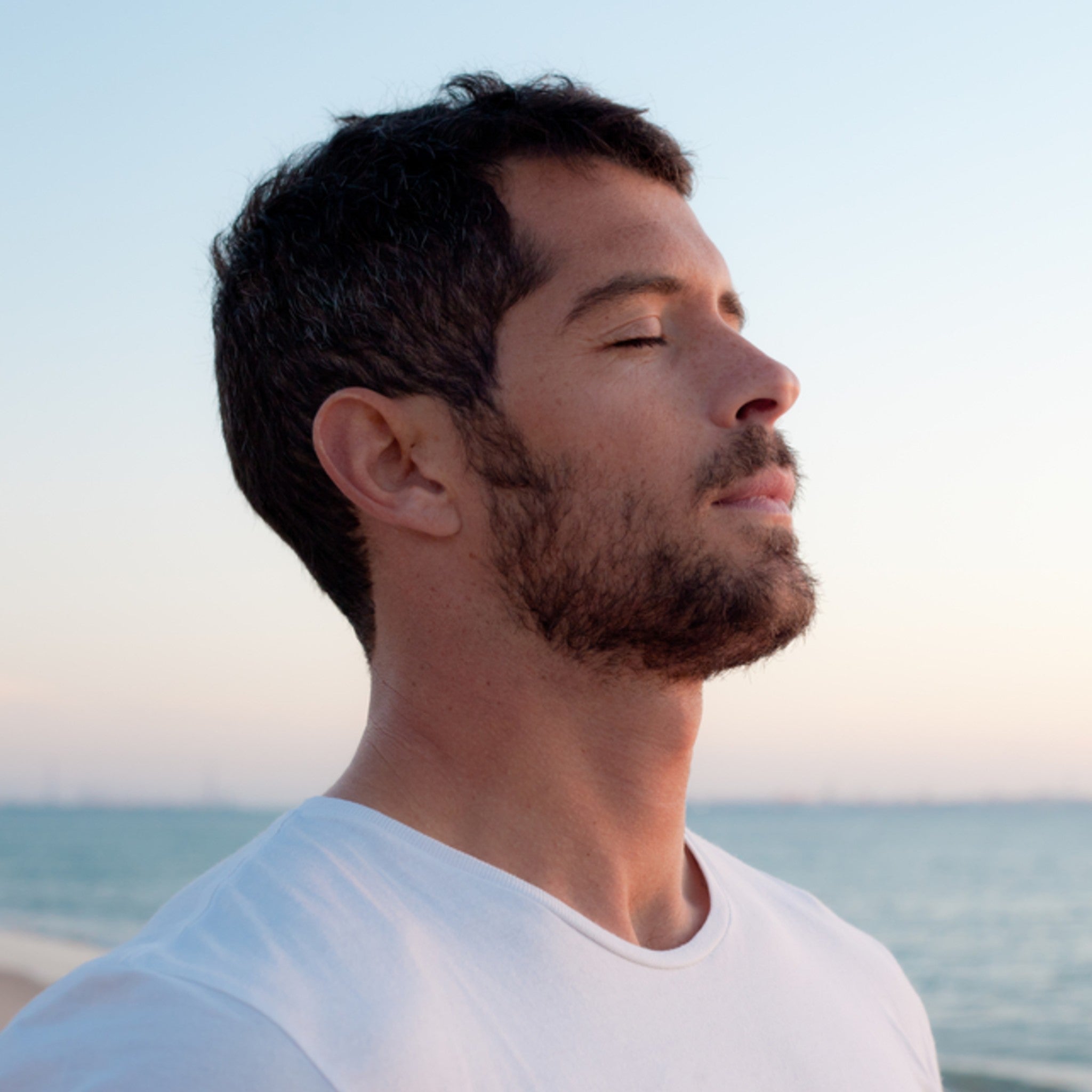 Ear Chakras Activation Maintenance Attunement: man with closed eyes and beard standing peacefully on a beach, facing the setting sun and ocean.