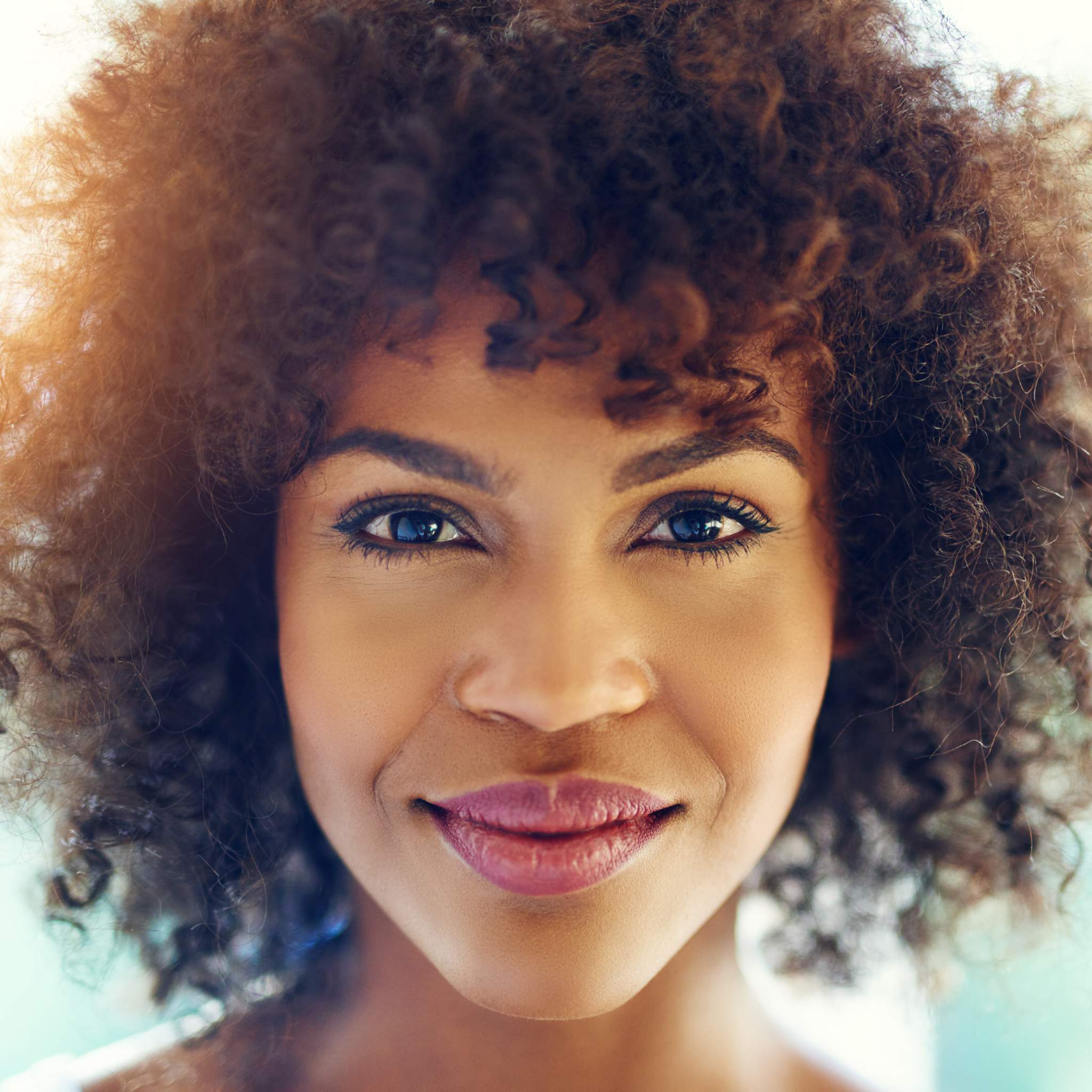 Close-up portrait of a woman with dark, curly hair smiling softly toward the camera, illuminated by warm sunlight.