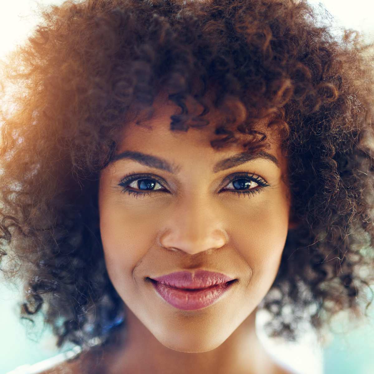Close-up portrait of a woman with dark, curly hair smiling softly toward the camera, illuminated by warm sunlight.