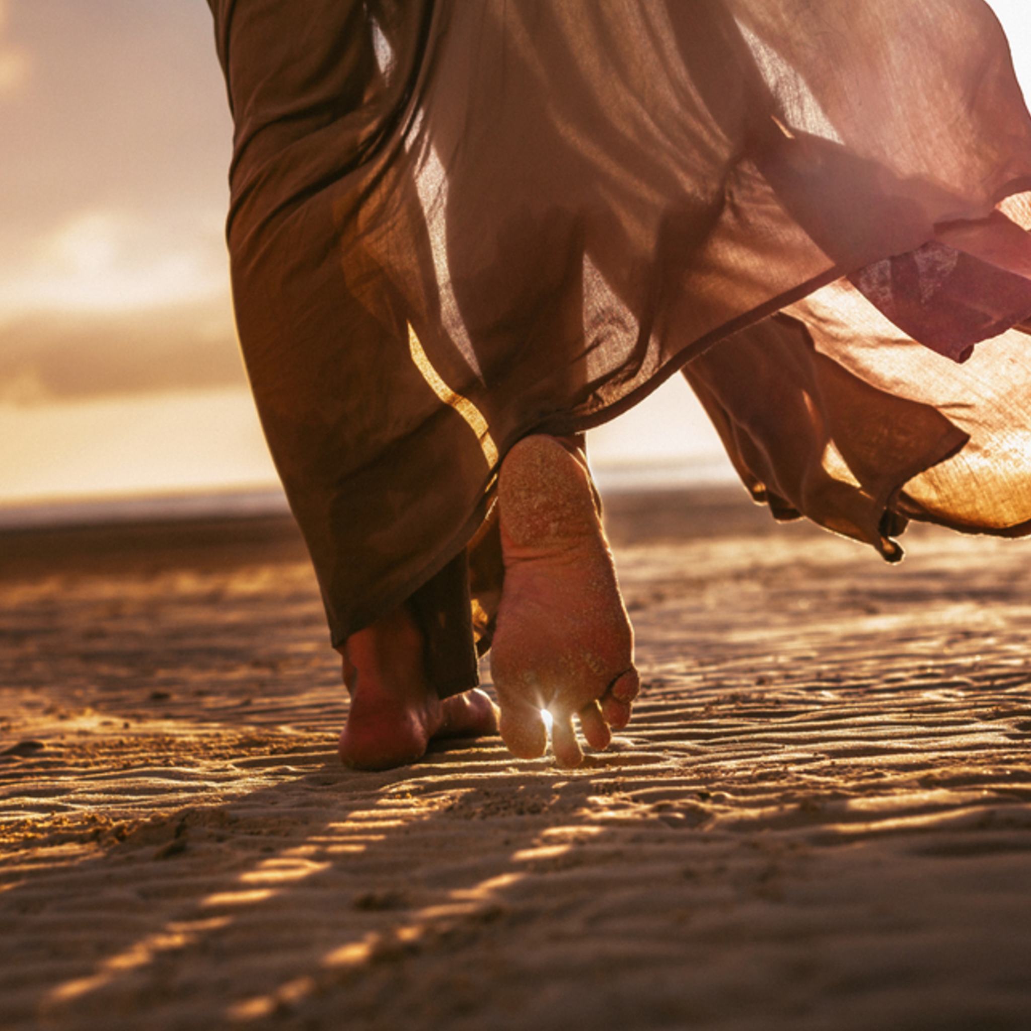 A low-angle, close-up shot of a person's bare feet walking across rippled sand on a beach at sunset, with a flowing, natural-coloured garment blowing in the wind barely visible above the ankles.