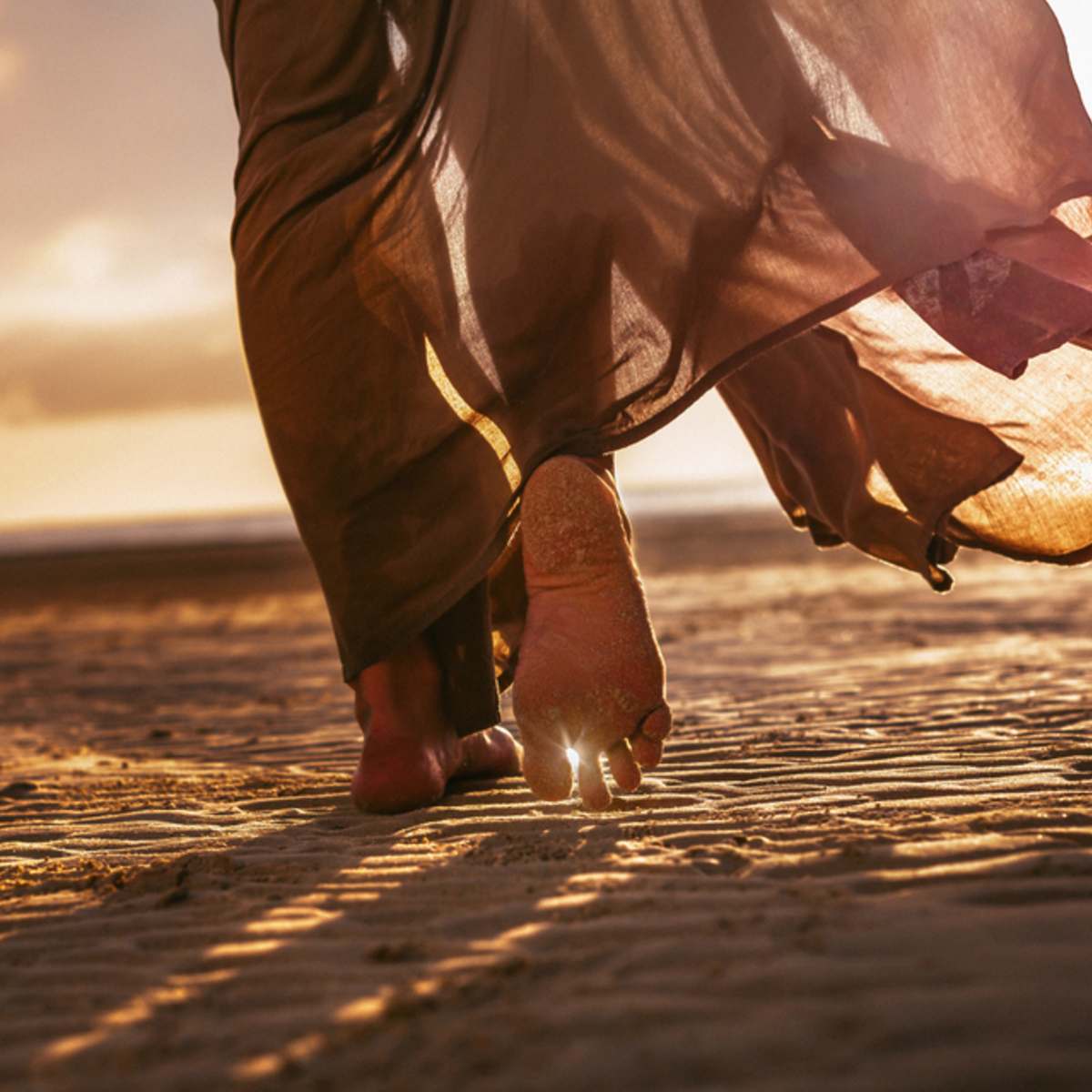 Person walking on a sandy beach with a long garment flowing behind them during sunset.