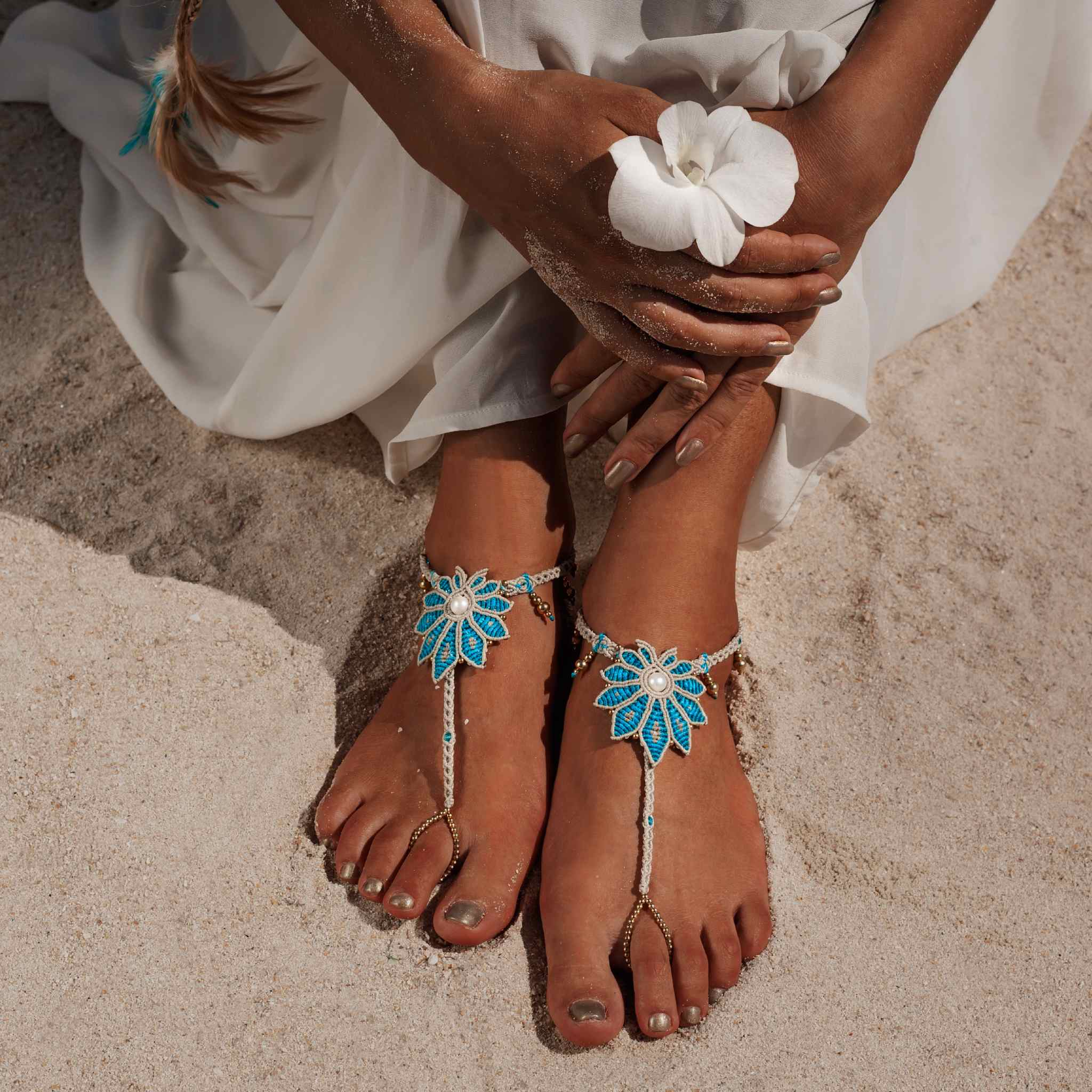 A close-up of a person's hands and feet, sitting cross-legged on light-coloured sand, wearing a flowing white garment. The feet are adorned with turquoise macrame barefoot sandals, and the hands gently hold a single white orchid or flower.