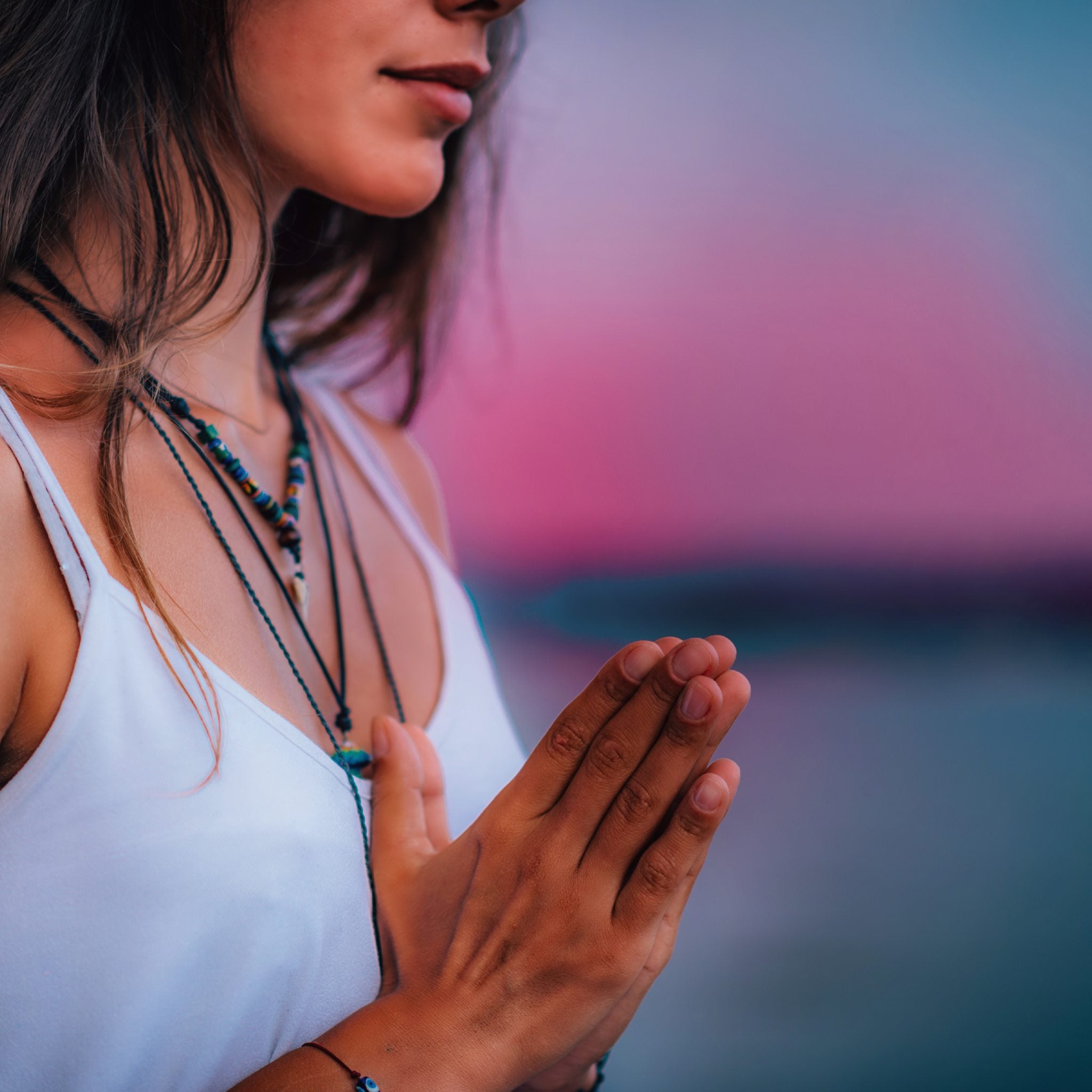 A close-up side profile of a woman meditating with her eyes closed and hands held in prayer position at her chest, set against a stunning, blurred background of vibrant pink, purple, and blue sunset over water.