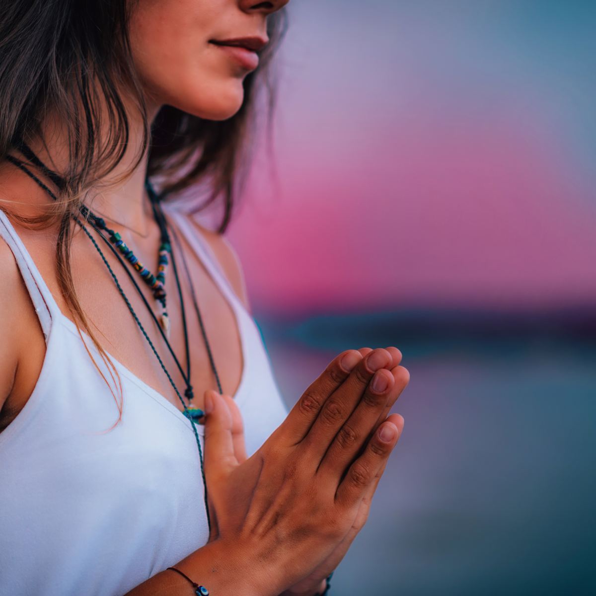 Woman meditating with hands held in prayer position set against a pink sunset.