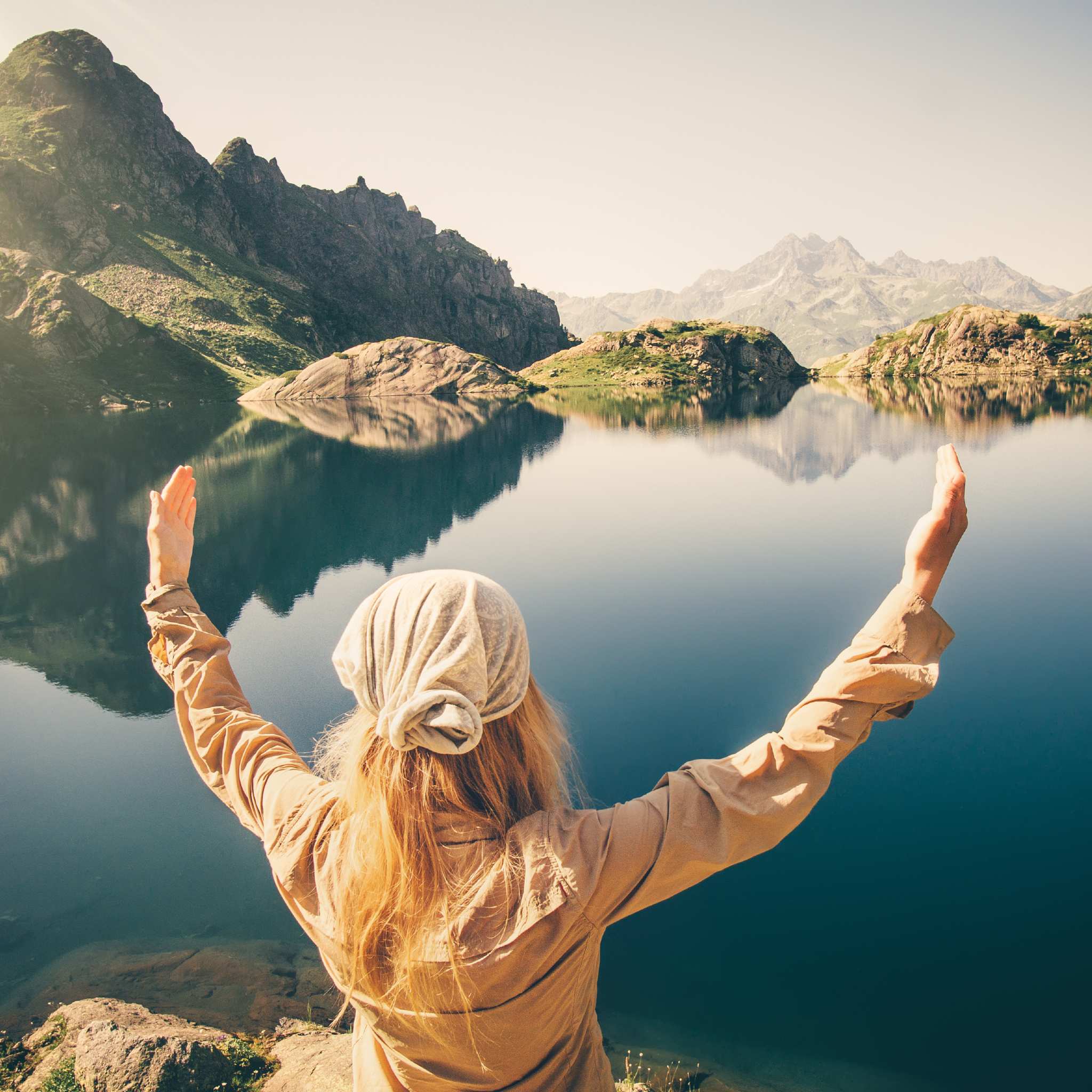 A person with long red hair and a bandana stands facing away, with arms raised to a reflective, calm blue alpine lake, surrounded by high, rocky mountains under a bright sky.