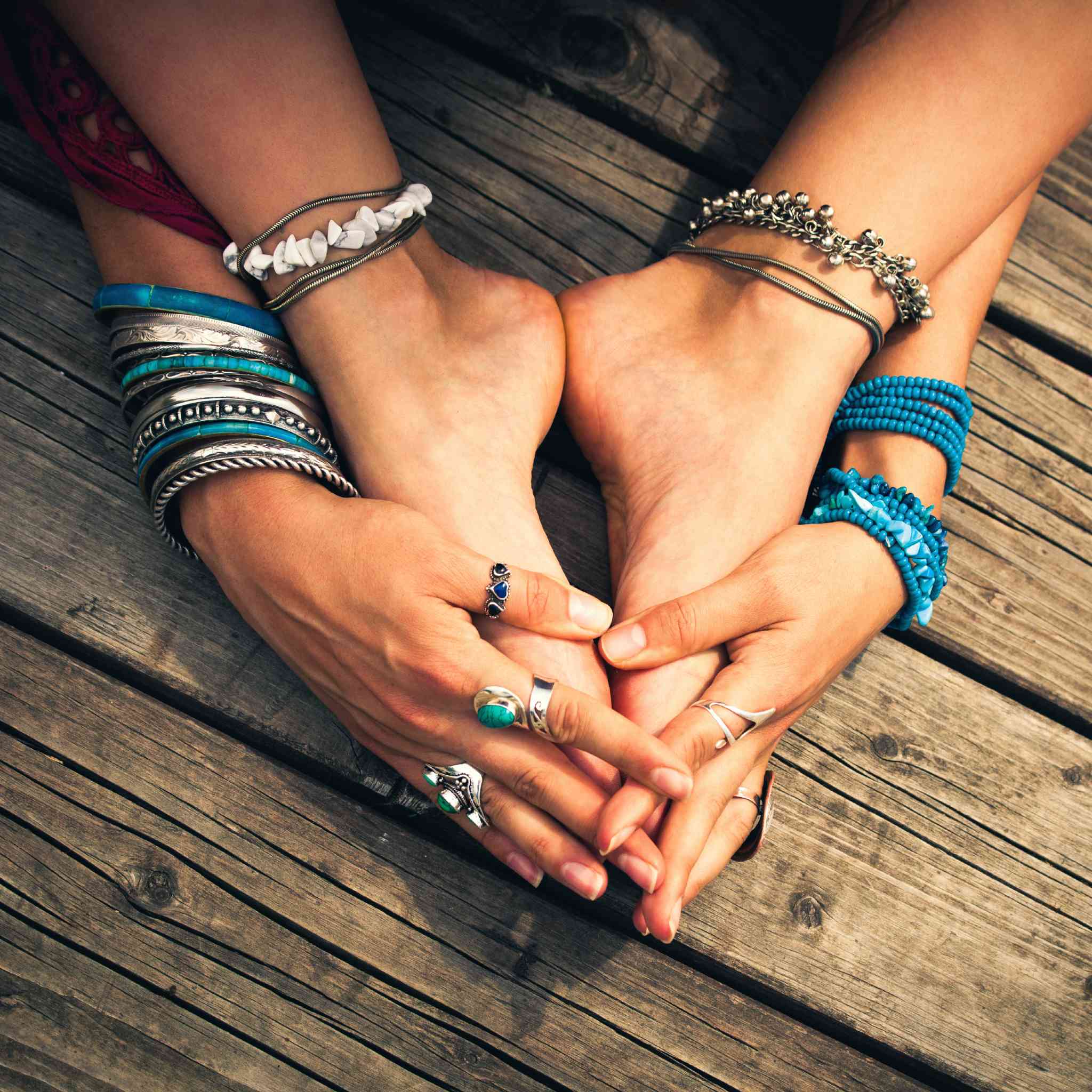A close-up of a person's bare feet and hands adorned with many silver and blue bracelets/rings, resting on wooden decking, signifying grounding.