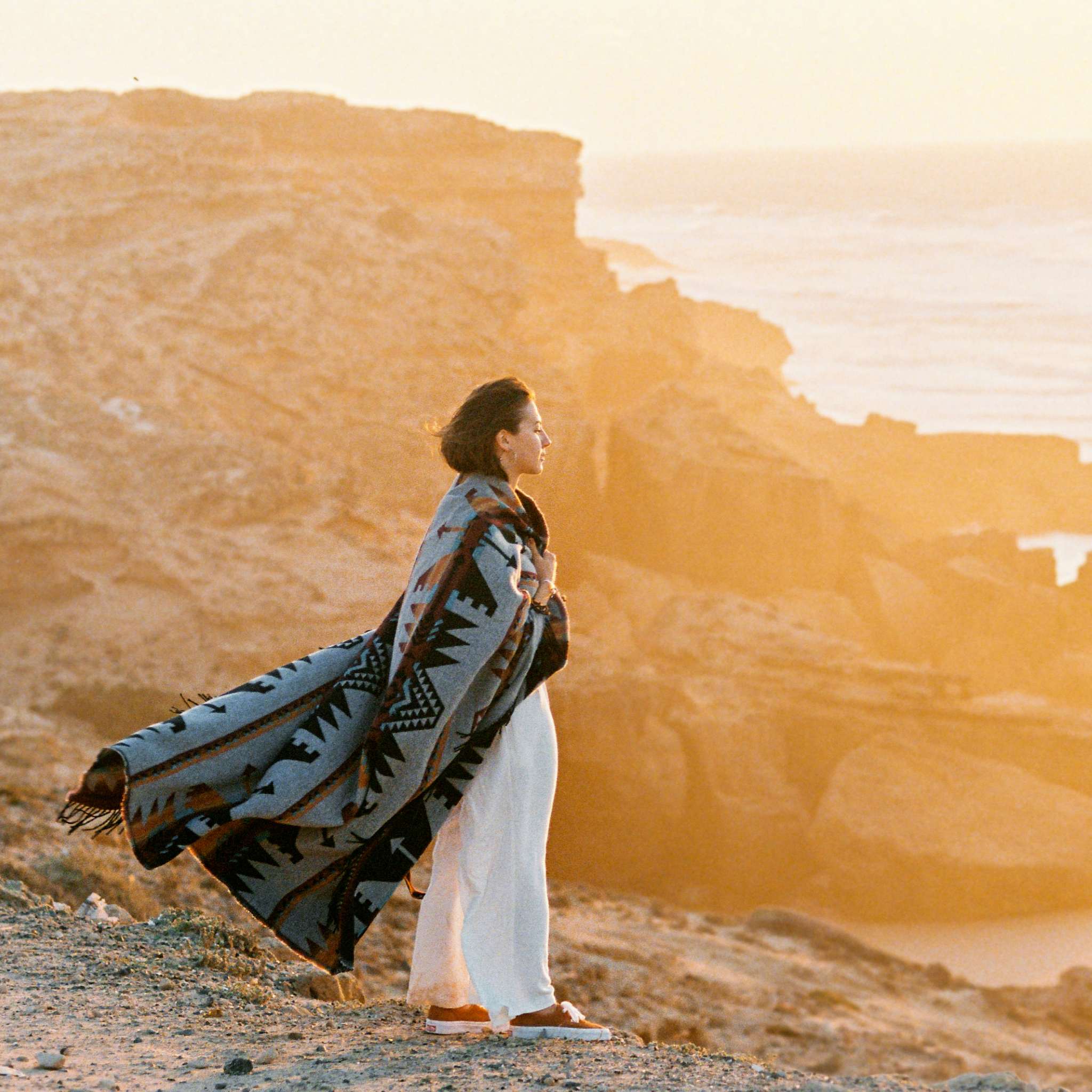 A woman wrapped in a patterned, southwestern-style blanket stands on a cliff overlooking the ocean, silhouetted by the warm, intense orange glow of sunset over a rugged landscape.