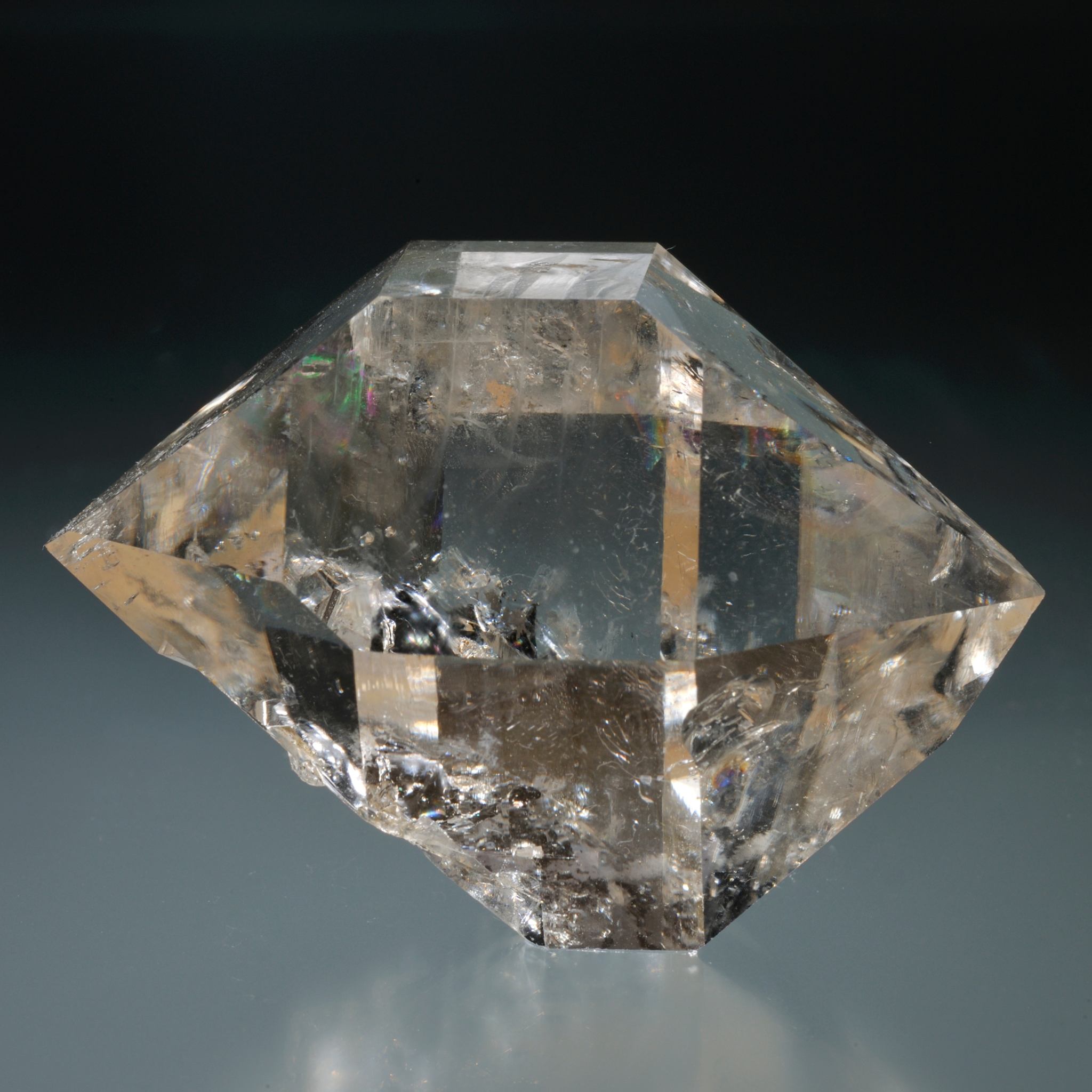 A close-up studio photograph of a double-terminated Herkimer Diamond quartz crystal. The clear, slightly golden stone shows many facets and glints of rainbow iridescence, set against a reflective grey surface and dark background.