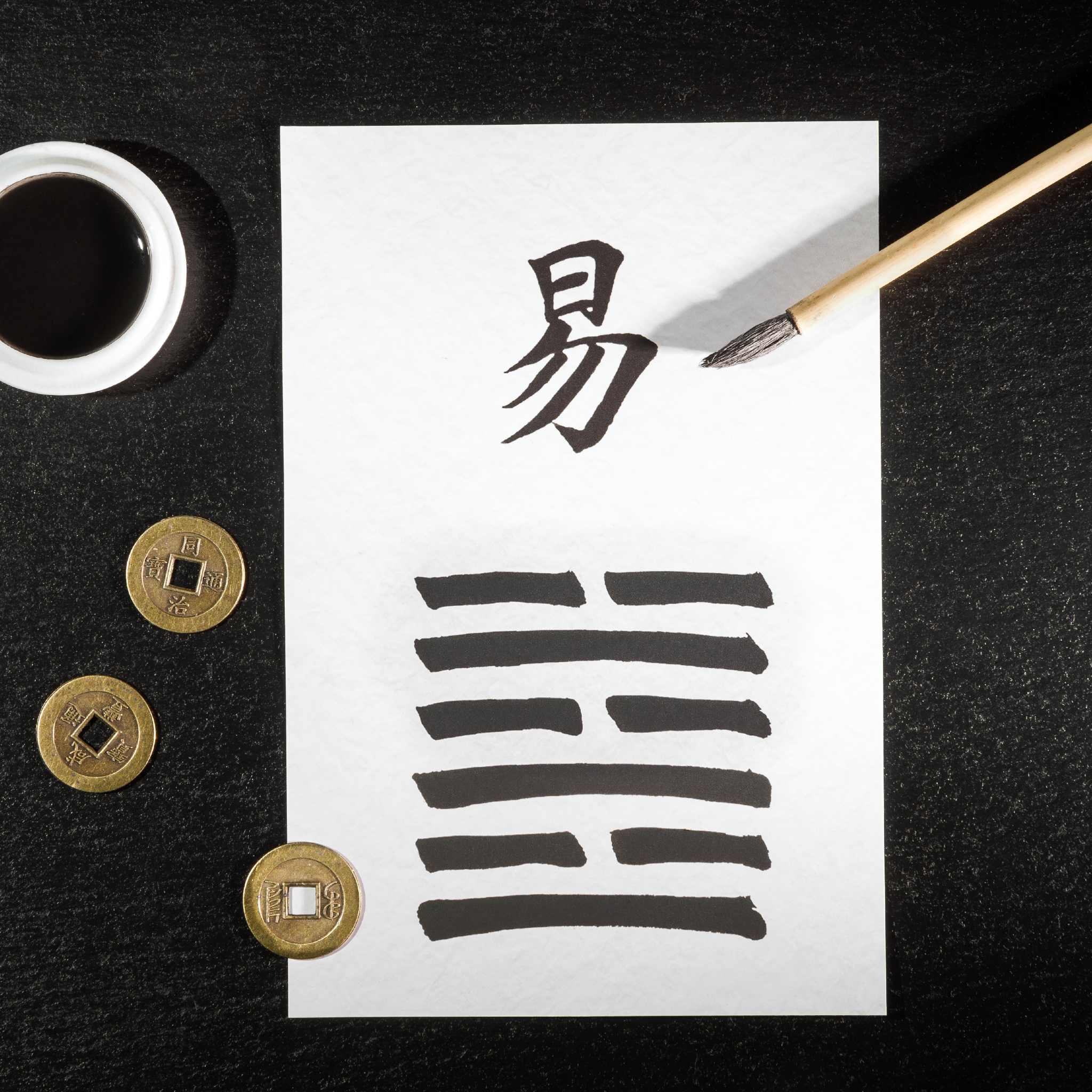 An overhead shot of a clean, dark surface featuring a white sheet of paper with black calligraphy. The paper displays the Chinese character for 'Yi' (meaning change or easy) above a six-line hexagram, representing the divination method of the I Ching. To the left are three traditional Chinese coins and a white dish of black ink, with a fine-tipped calligraphy brush on the right, signifying the focus of the I Ching path alignment dedication.