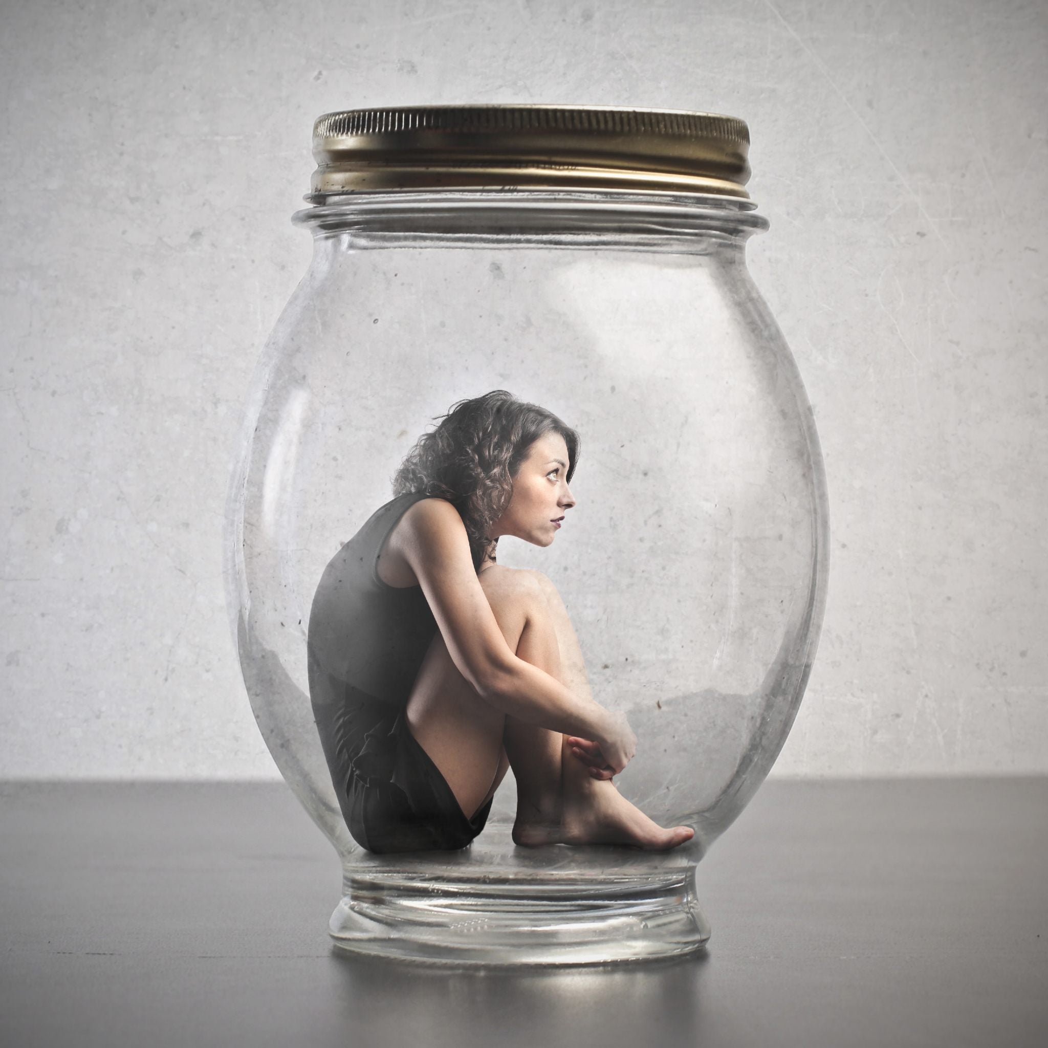 Person sitting inside a glass jar with a metal lid on a neutral background represents inappropriate fear.
