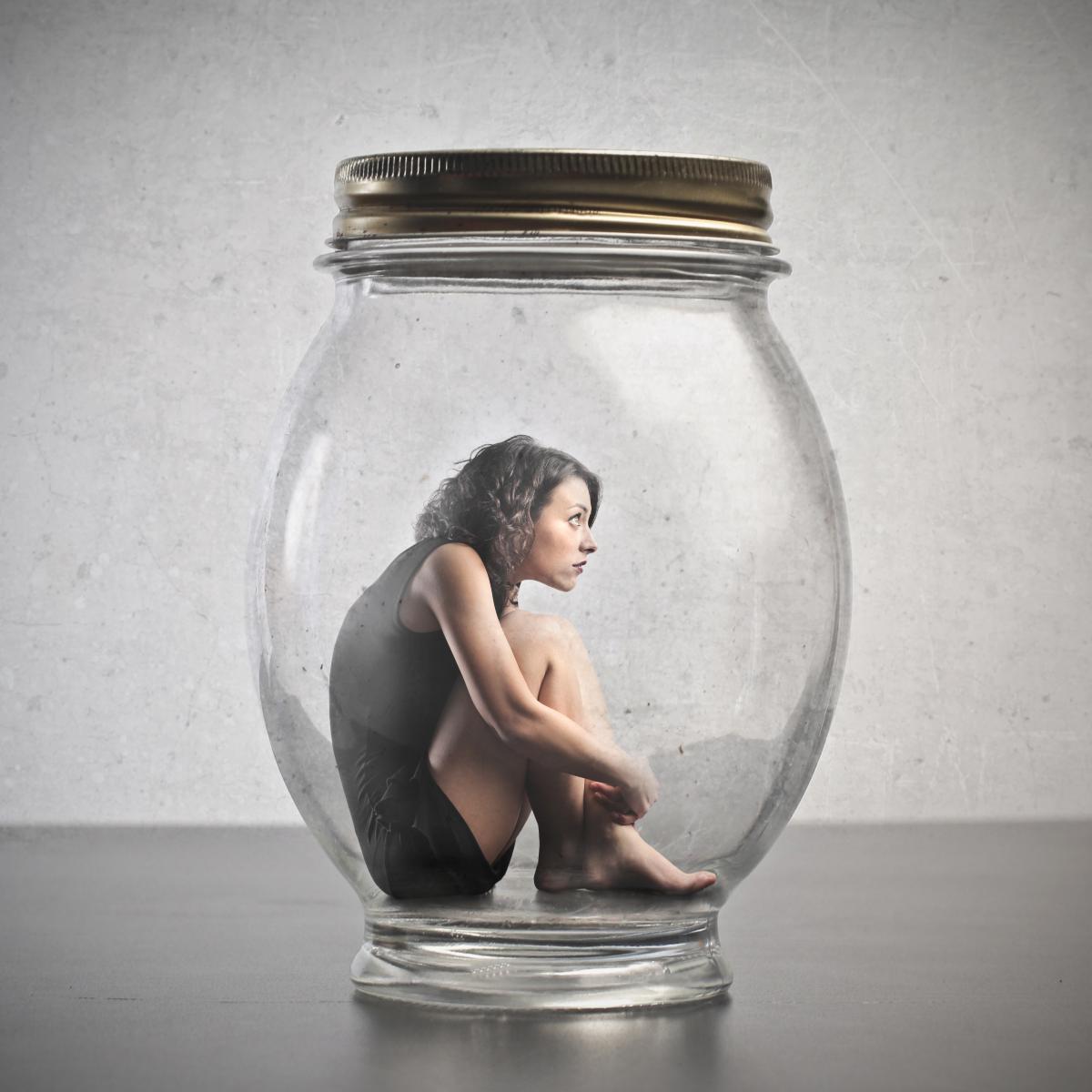 Person sitting inside a glass jar with a metal lid on a neutral background represents inappropriate fear.