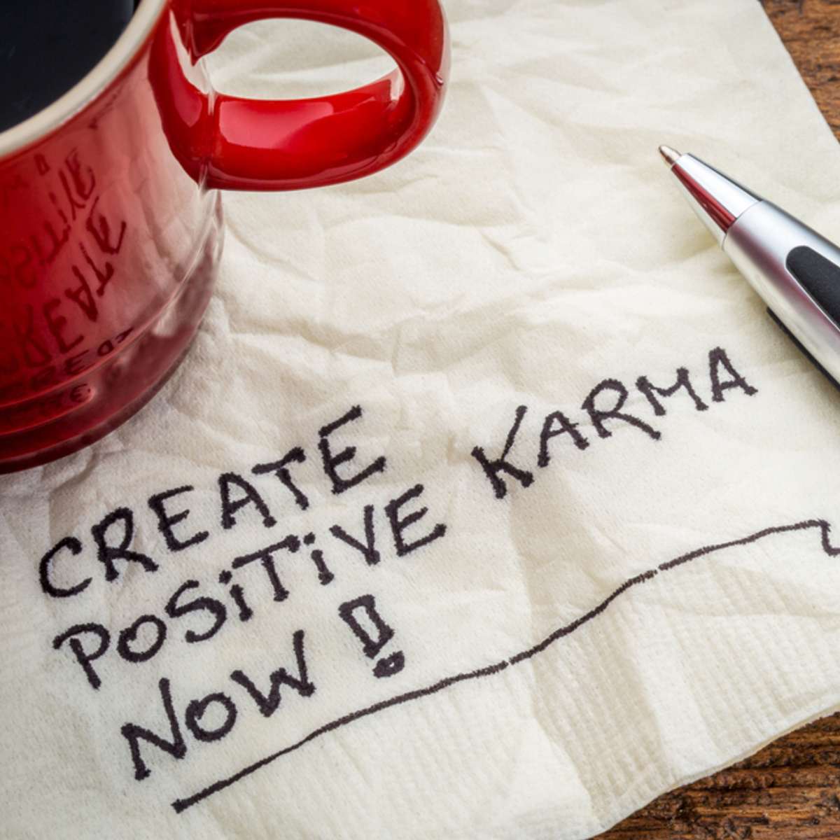 Red mug, pen, and napkin with 'Create Positive Karma Now!' text on a wooden surface.