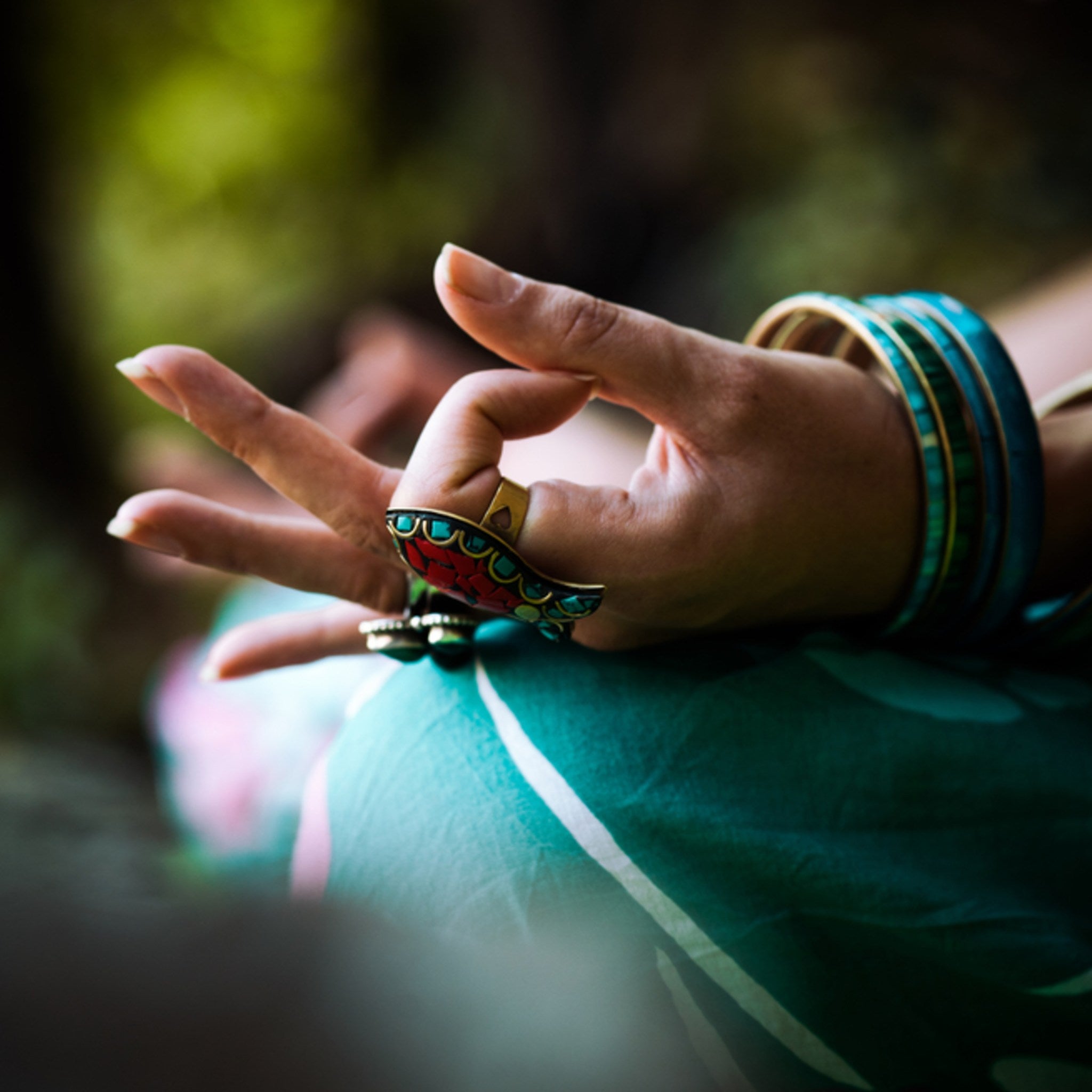 Close-up of a hand in a mudra (yoga hand position), wearing vibrant turquoise bangles and a large red-and-turquoise ring, resting on a green patterned lap.