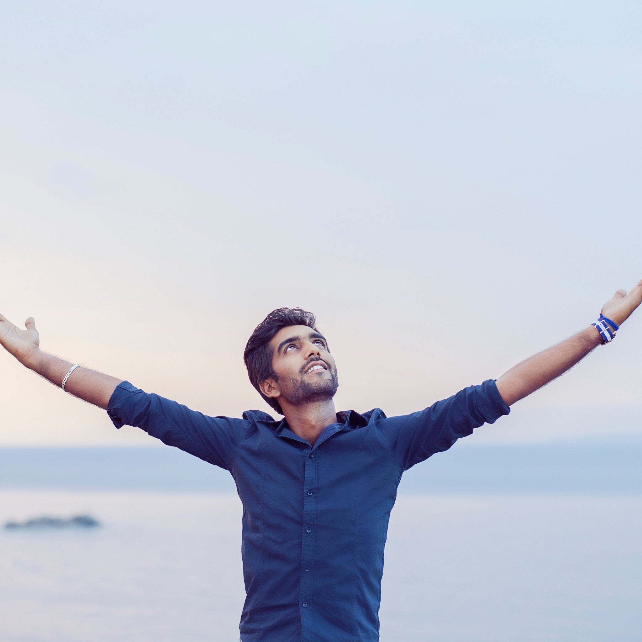 A young man wearing a dark button-down shirt stands outdoors, facing forward with his eyes and head tilted upward, arms outstretched and palms open towards the sky against a soft blue and pink horizon over water.